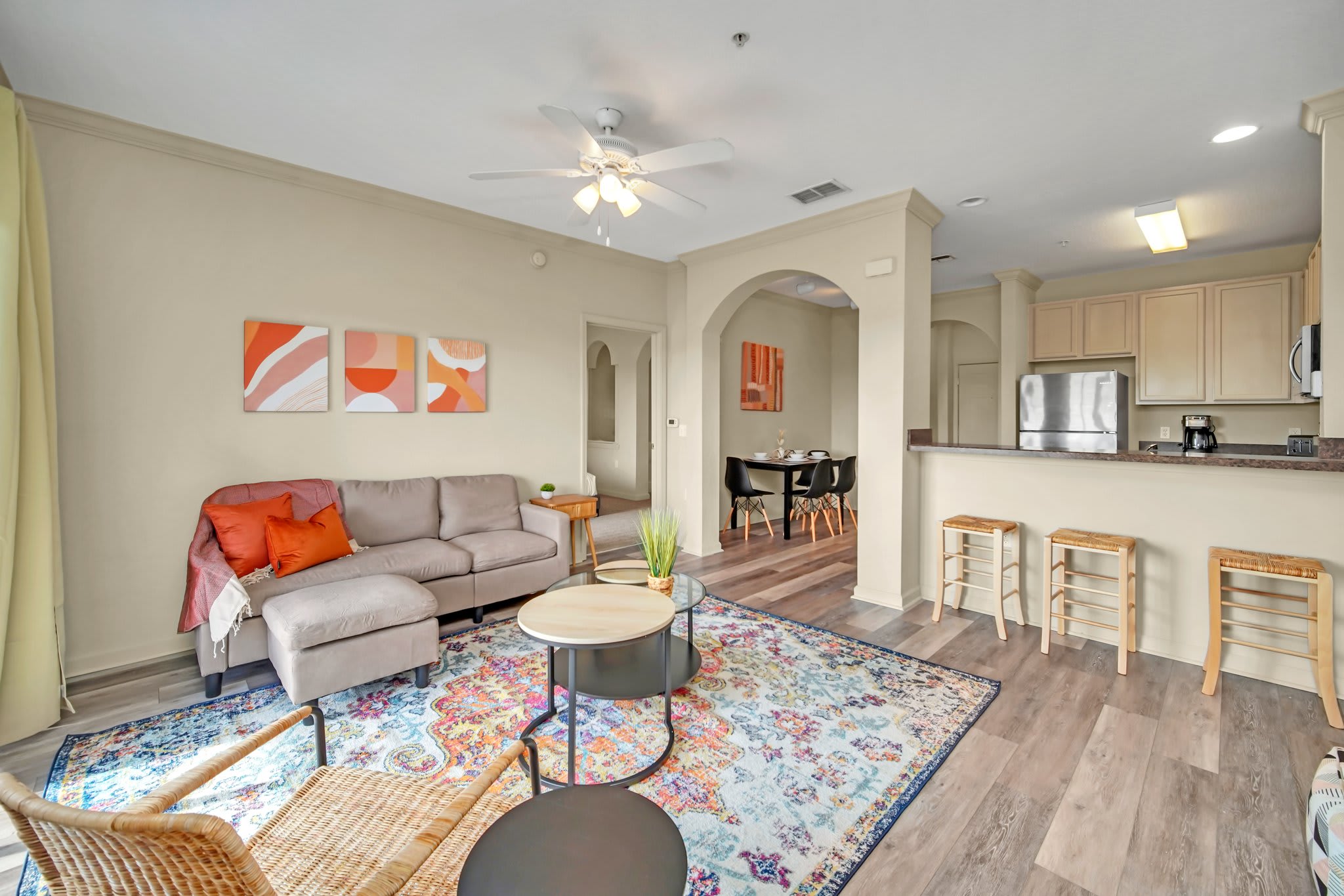 The living room is furnished with a light gray sectional sofa, a colorful area rug, and a set of nesting coffee tables. Abstract art adorns the wall, and a ceiling fan provides comfort.