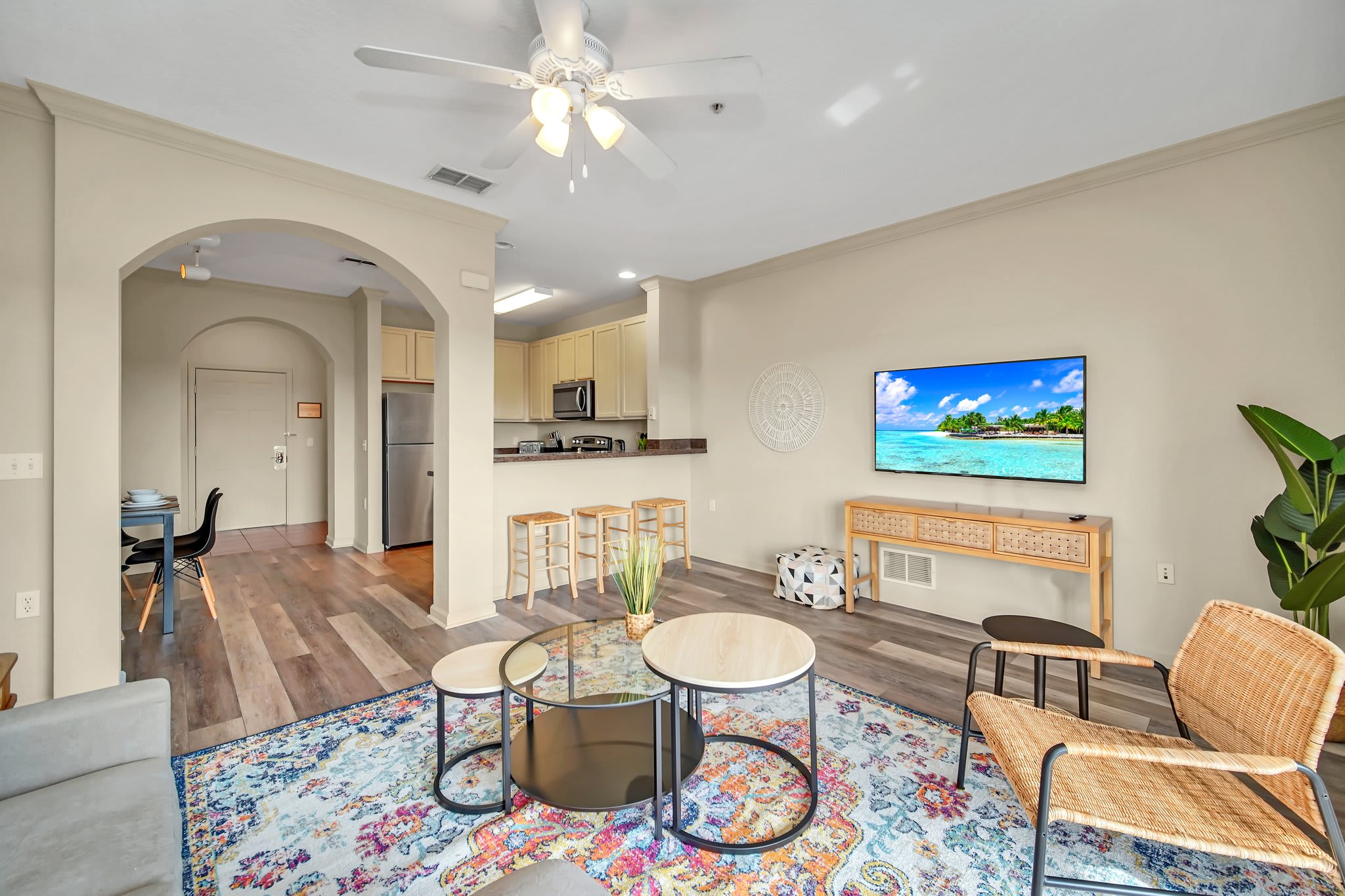 An open-concept living space with a light gray sofa, a vibrant patterned rug, and a view into the kitchen with a breakfast bar. A large flat-screen TV is mounted on a console, and a ceiling fan hangs overhead.