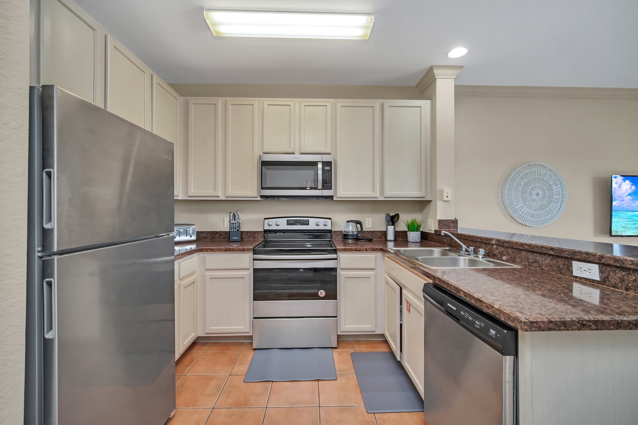 A well-equipped kitchen features light beige cabinetry, dark countertops, and stainless steel appliances including a refrigerator, microwave, oven, and dishwasher. Terracotta-colored tile flooring completes the space.