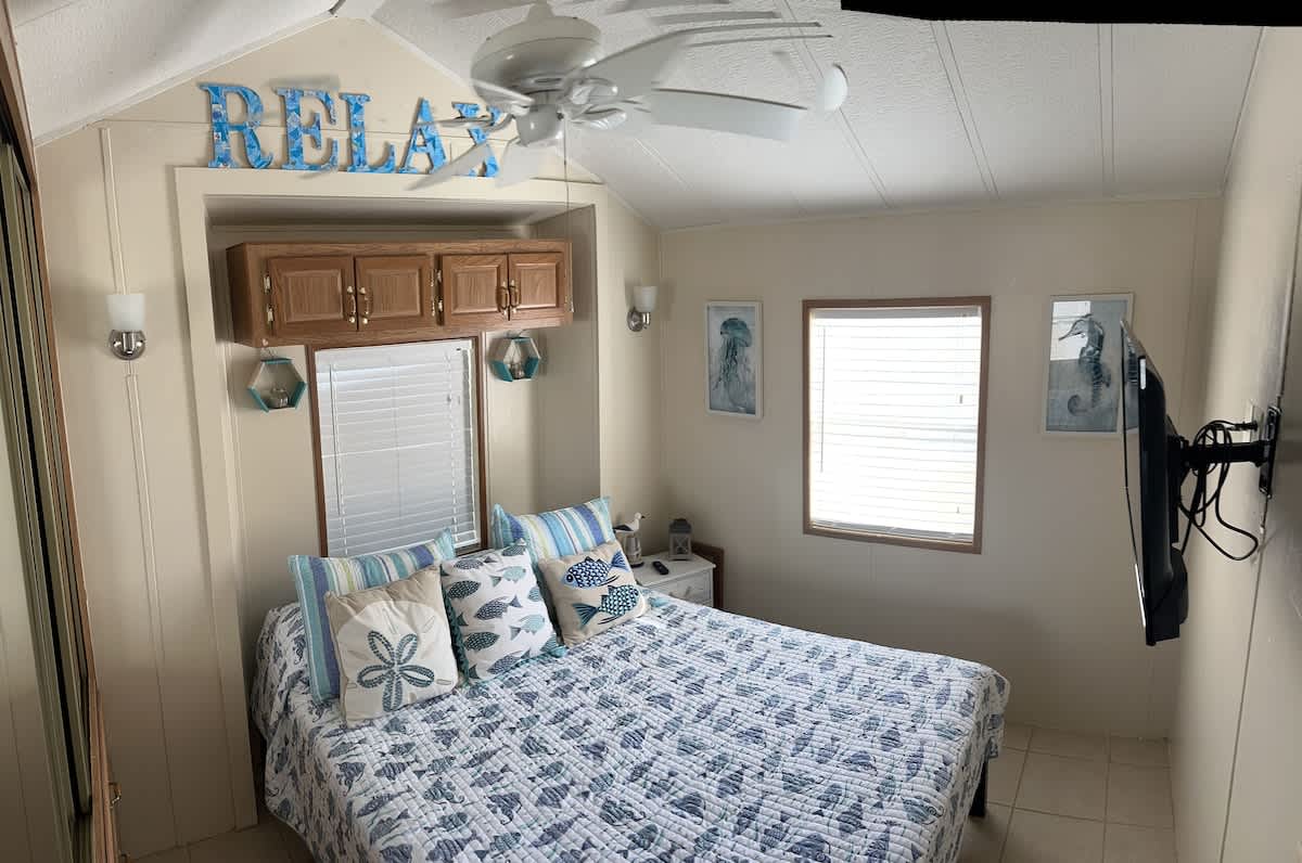 A cozy bedroom with a queen-sized bed featuring a blue and white fish-patterned quilt and decorative pillows. The room has light walls, a sloped ceiling with a fan, and coastal-themed decor including a "RELAX" sign and framed artwork.