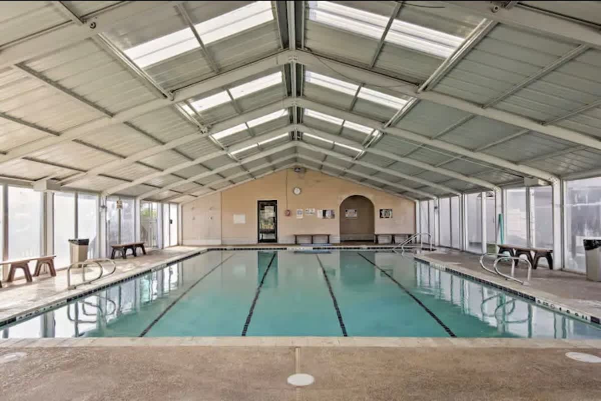 A long, rectangular indoor swimming pool with lane lines, housed under a high, vaulted metal roof with skylights, surrounded by light-colored walls and benches.