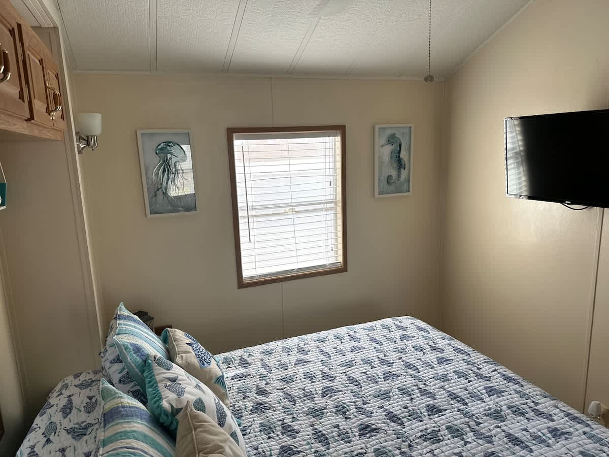 Another view of the cozy bedroom, highlighting the queen bed with its blue and white fish-patterned quilt. Two framed coastal artworks, one of a jellyfish and one of a seahorse, adorn the light-colored walls around a window with blinds.