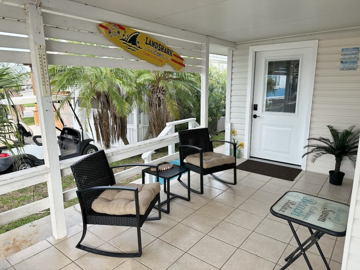 Covered outdoor patio with light-colored tiled floor, white wooden railing, two black metal rocking chairs with beige cushions, a small glass-top side table, and a yellow "Landshark" surfboard decoration.