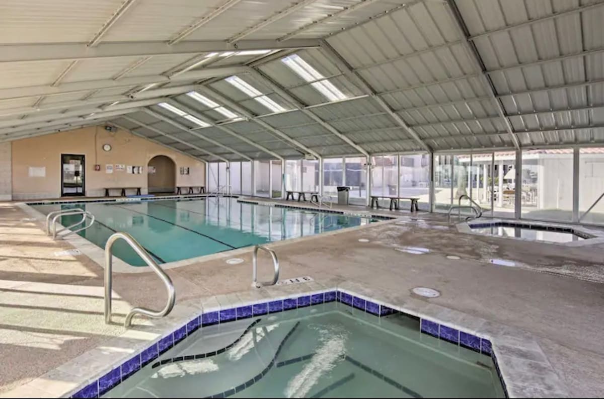 A spacious indoor pool area featuring a rectangular swimming pool with lane lines and an adjacent square hot tub with blue mosaic tiles, all under a high, skylit metal roof.