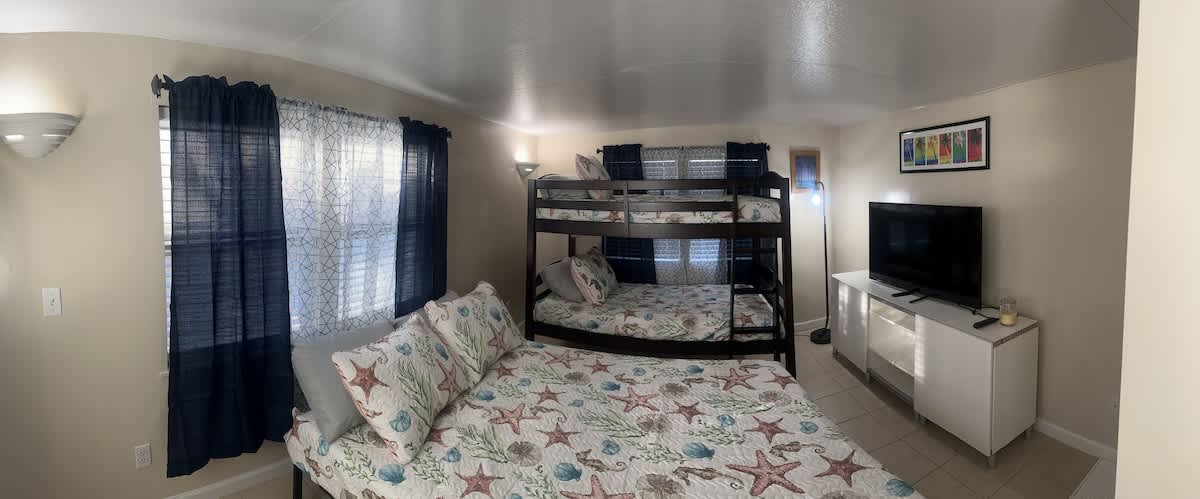 A wider view of the family-friendly bedroom, showing the queen bed and bunk beds with matching starfish-patterned bedding. Dark blue curtains frame the windows, and coastal artwork adorns the light-colored walls.