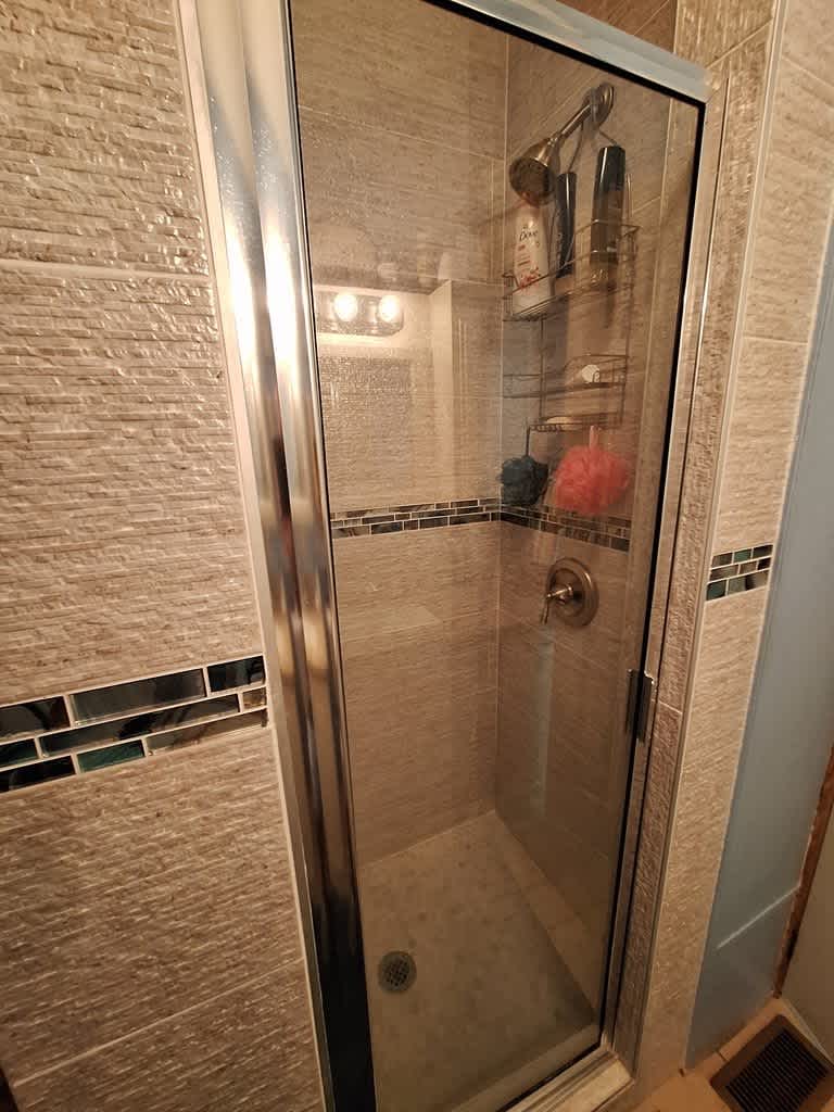 A close-up view of the modern walk-in shower with light-colored textured tile walls and a decorative blue and green mosaic accent strip. The shower features a glass door and a built-in shelf for toiletries.