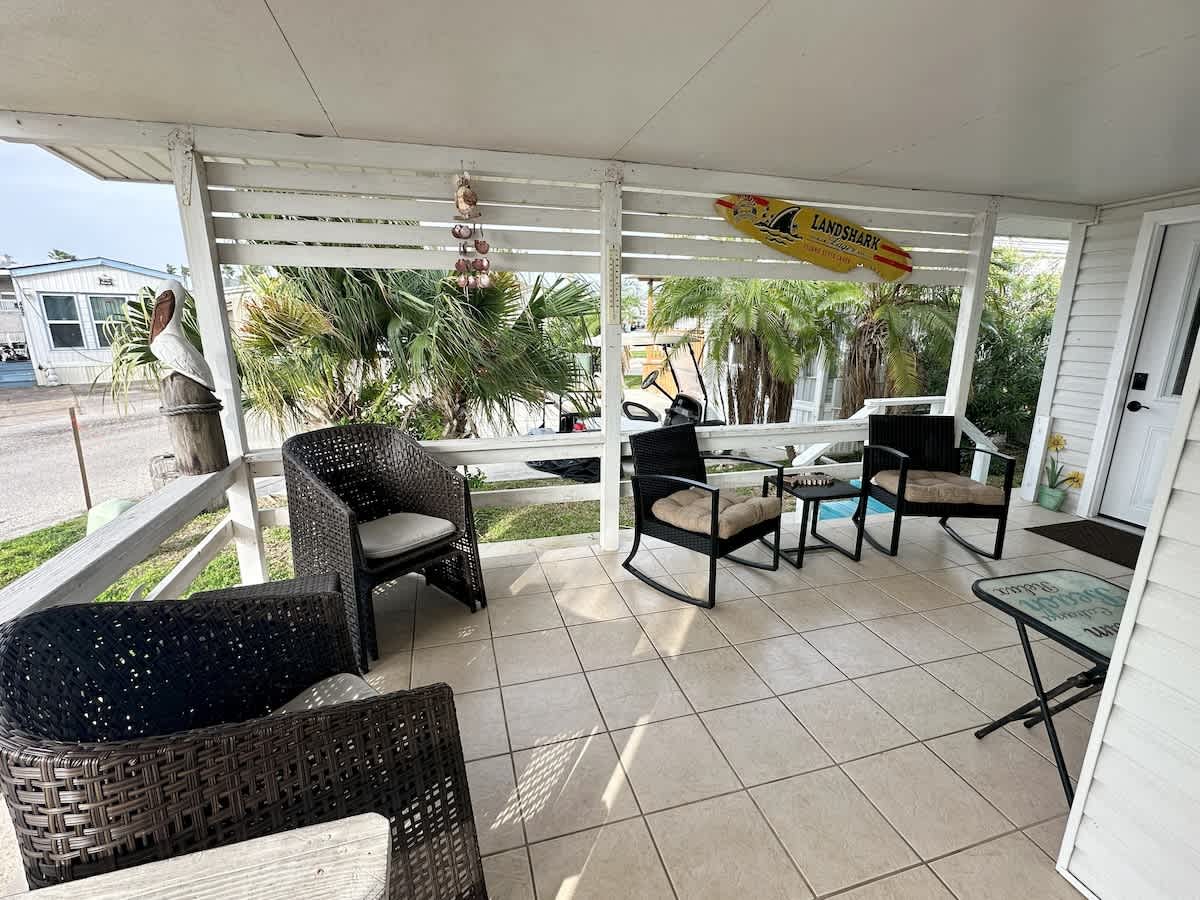 Covered outdoor patio with light-colored tiled floor, white wooden railing, a mix of dark wicker and black metal outdoor seating, and coastal decorations including a surfboard and shell wind chimes.