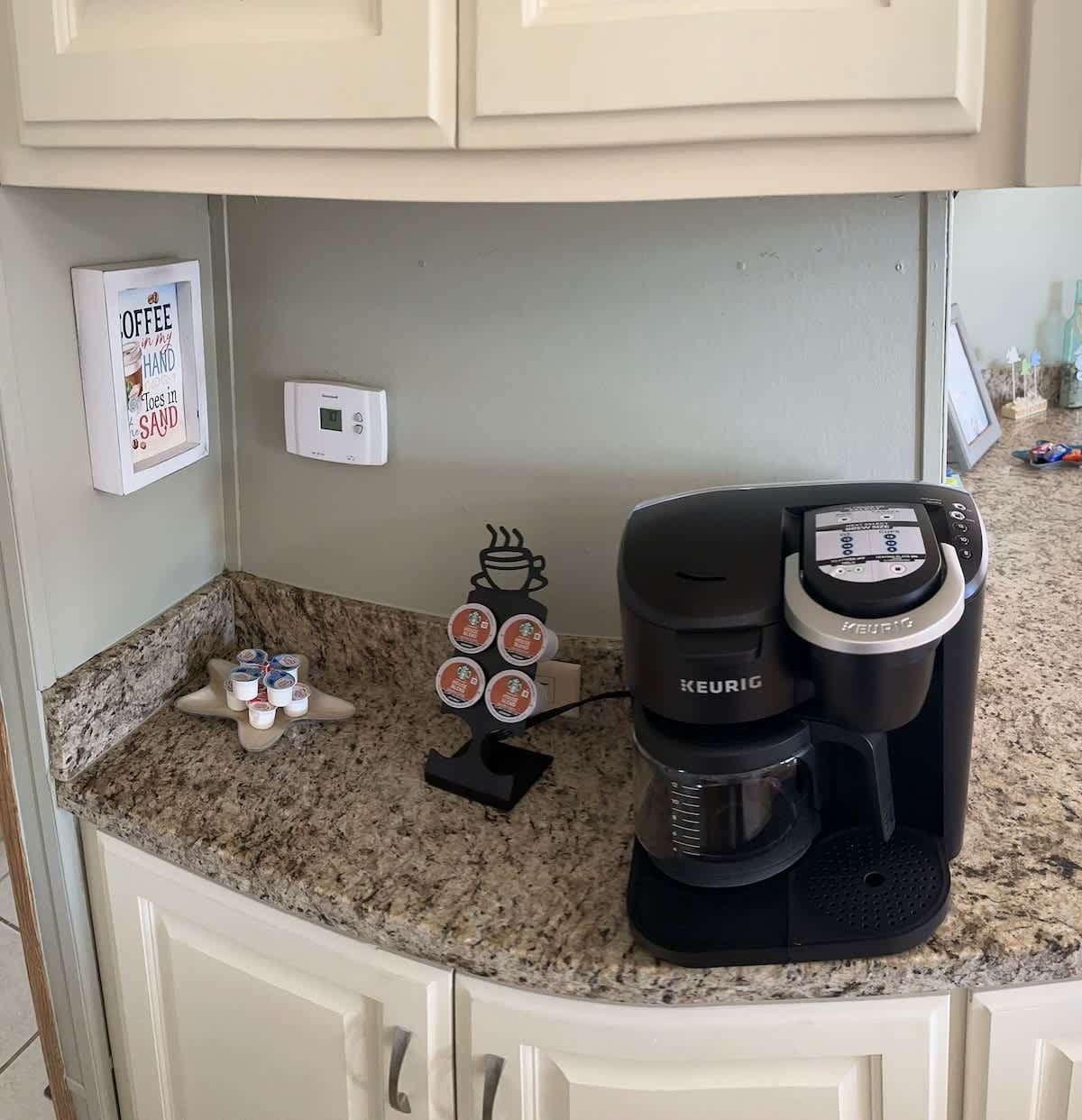 A dedicated coffee station in the kitchen featuring a black Keurig coffee maker, a K-cup pod holder, and a small framed sign. The area has light-colored walls and granite-look countertops.