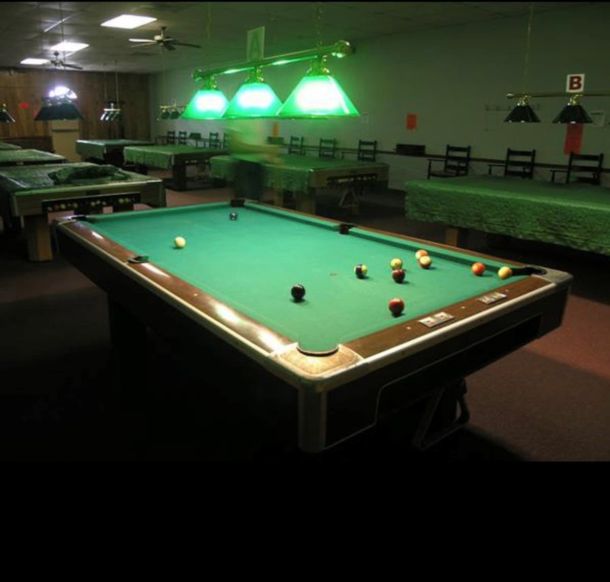 A communal game room featuring multiple green-felted pool tables under bright green overhead lamps, suggesting a recreational area within the resort. The room has dark flooring and simple chairs.