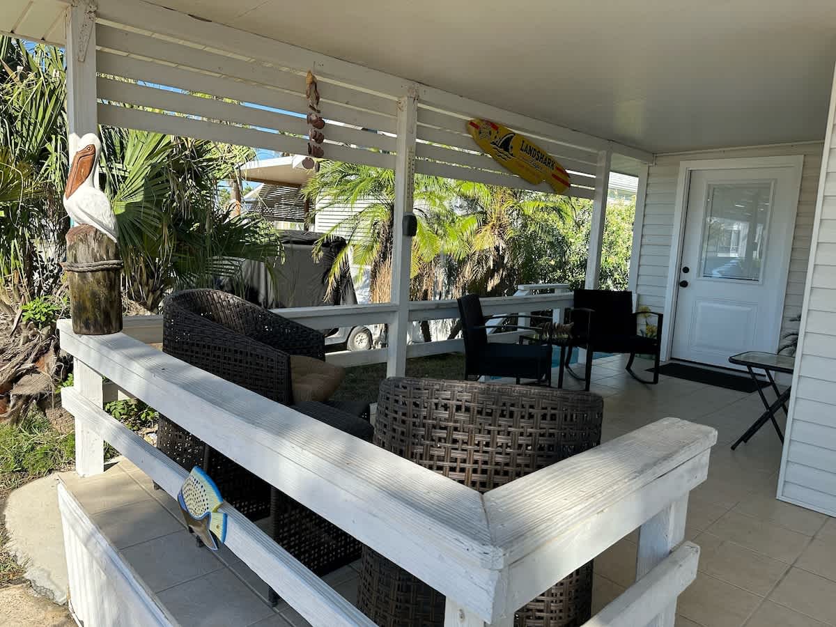 Wide view of a covered outdoor patio with light-colored tiled floor, white wooden railing, dark wicker seating, a wooden pelican statue, and a "Landshark" surfboard decoration, with lush palm trees in the background.