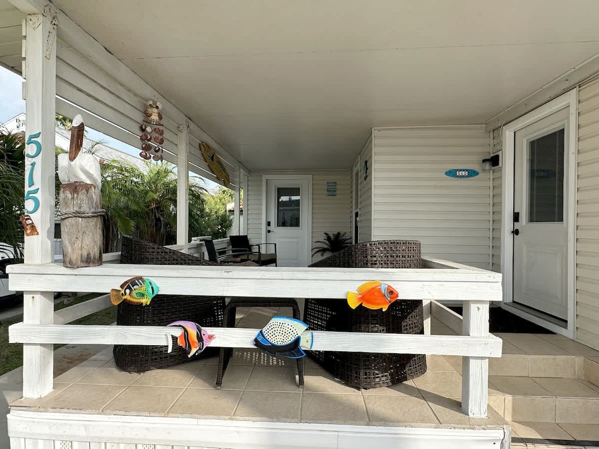 Covered outdoor patio featuring a white wooden railing decorated with colorful fish accents, dark wicker seating, and a prominent wooden pelican statue on a post, all on a tiled floor.