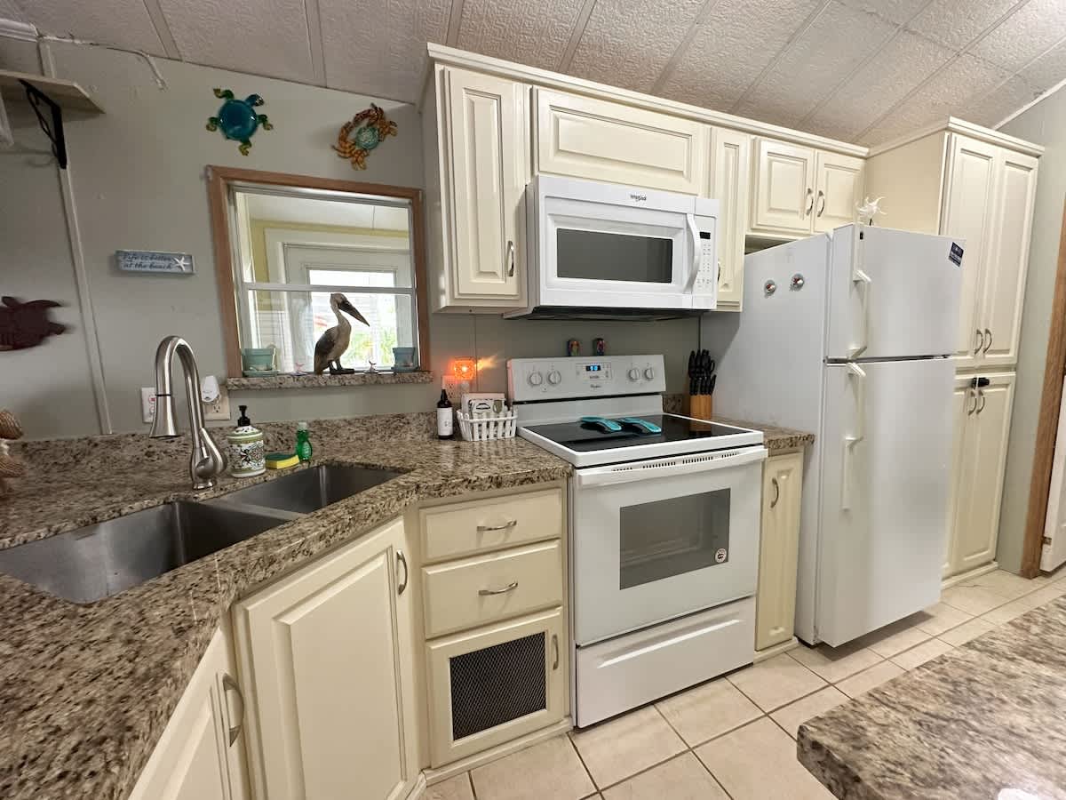 A bright kitchen with cream-colored cabinets, granite-look countertops, a double stainless steel sink, and white appliances including a range, microwave, and refrigerator. Coastal decor like a pelican statue and turtle wall art is visible.