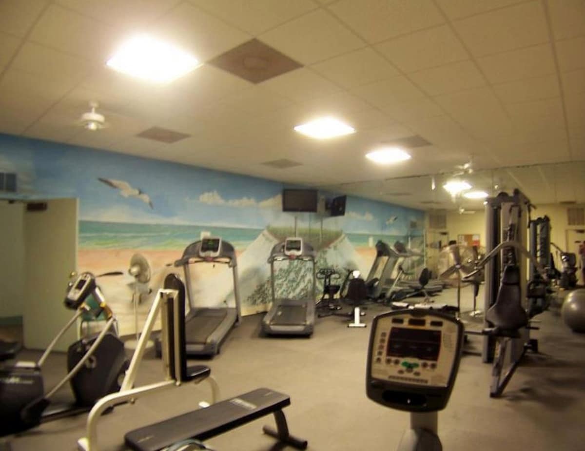 A well-equipped fitness room featuring various exercise machines including treadmills, ellipticals, and weight machines, set against a vibrant mural depicting a beach scene.