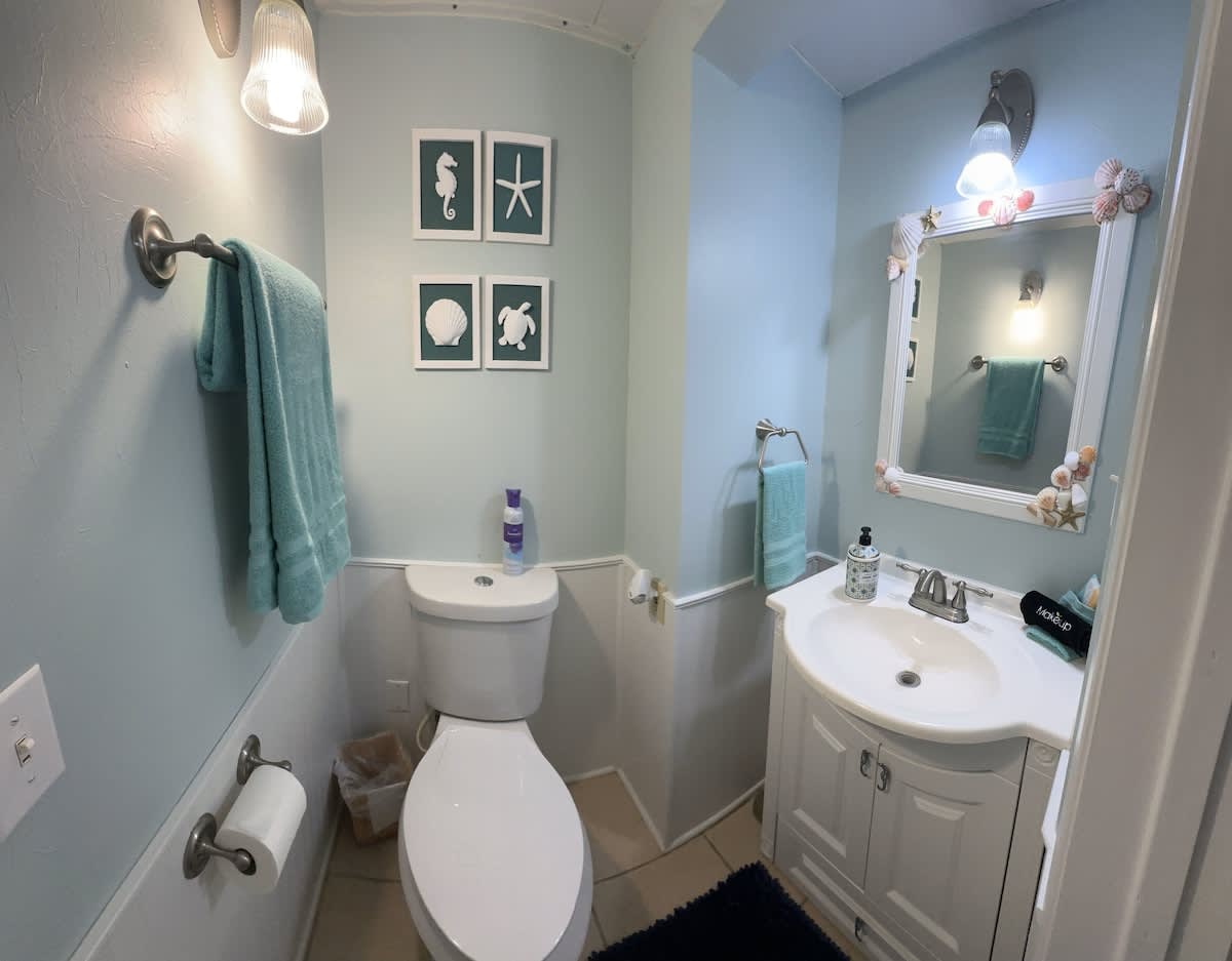 A half bathroom with light blue walls and white wainscoting, featuring a corner vanity with a white sink and a toilet. Coastal decor, including seashell-framed mirror and framed sea-themed art, enhances the beachy ambiance.