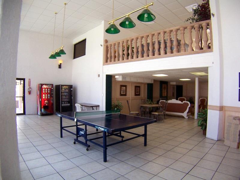A bright communal game room with white tiled floors, featuring a blue ping-pong table in the foreground, vending machines, and a lounge area with a sofa visible through an archway.