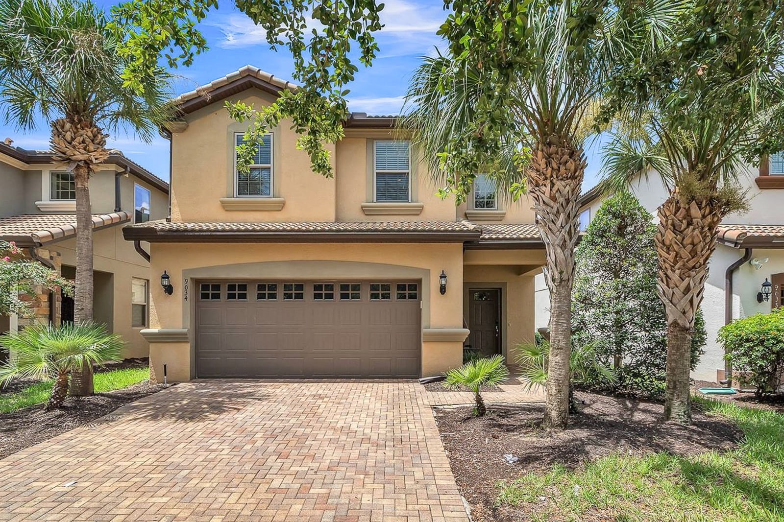 The front exterior of a two-story, light tan house with a brown garage door and a matching front door. A brick paver driveway leads to the garage, flanked by several palm trees and green landscaping under a clear blue sky.