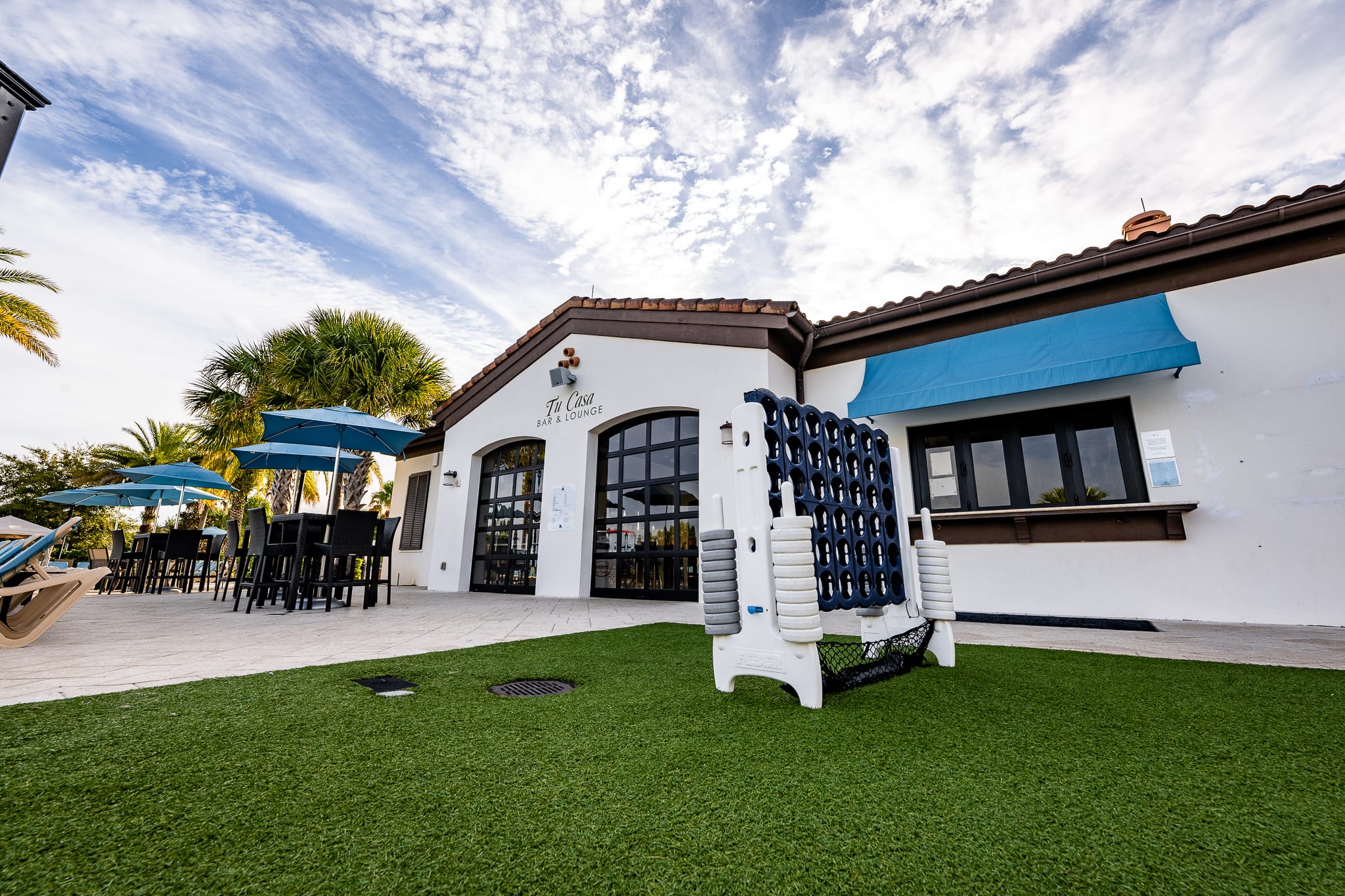 An outdoor bar and lounge area with tables and chairs under blue umbrellas, adjacent to a building with 'The Cove Bar & Lounge' signage. A large outdoor Connect Four game is prominently featured on artificial turf.