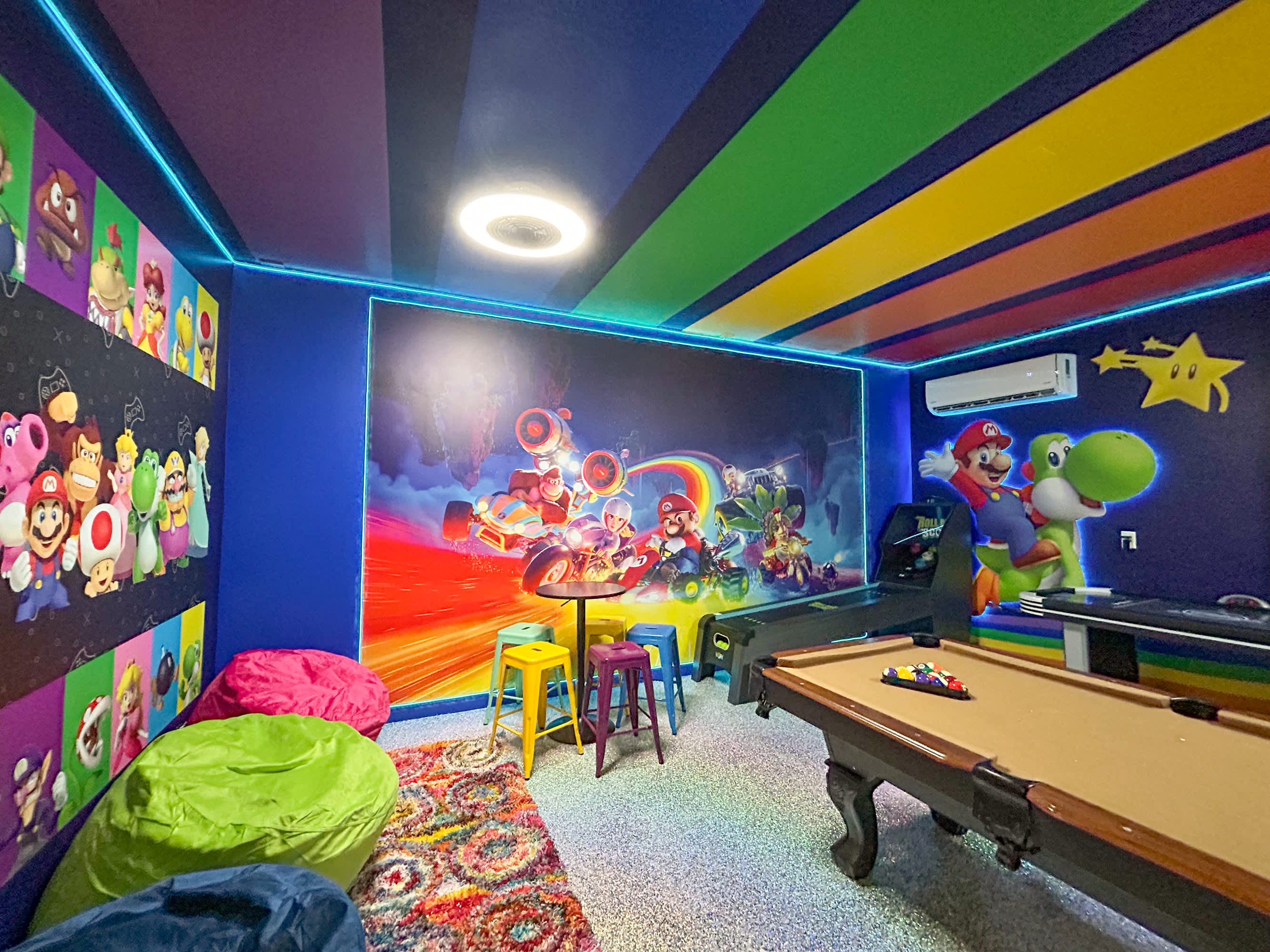 A vibrant game room with a pool table, bean bag chairs, and a small round table with colorful stools, all set against walls with Mario-themed murals and neon lighting. The ceiling is painted with colorful stripes.