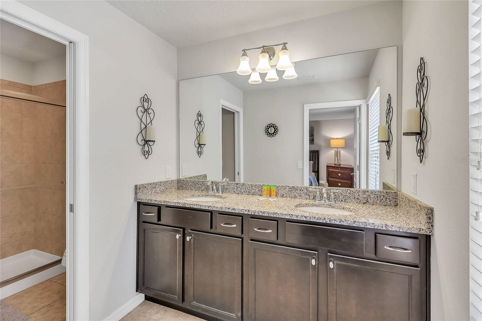 A modern bathroom with a double vanity featuring a granite countertop, two undermount sinks, and dark wood cabinetry. A large mirror spans the width of the vanity, reflecting a shower with tiled walls and a toilet in an adjacent room.