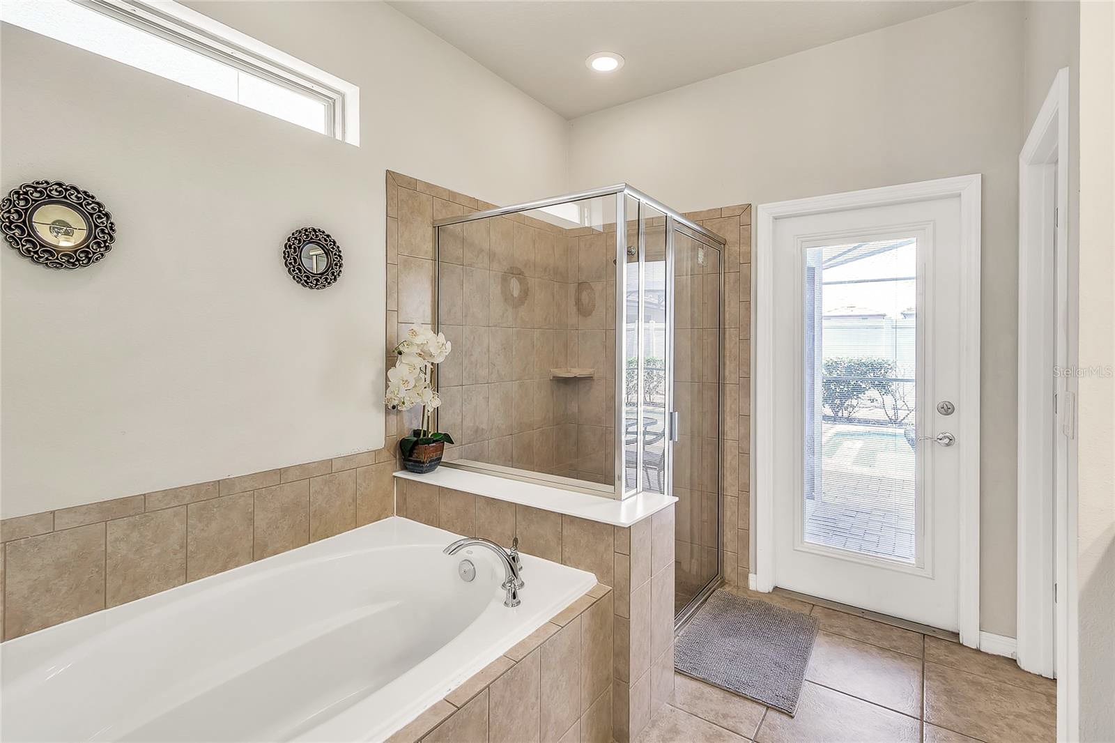 A spacious bathroom featuring a large built-in bathtub, a glass-enclosed walk-in shower with tiled walls, and a door providing direct access to an outdoor space.