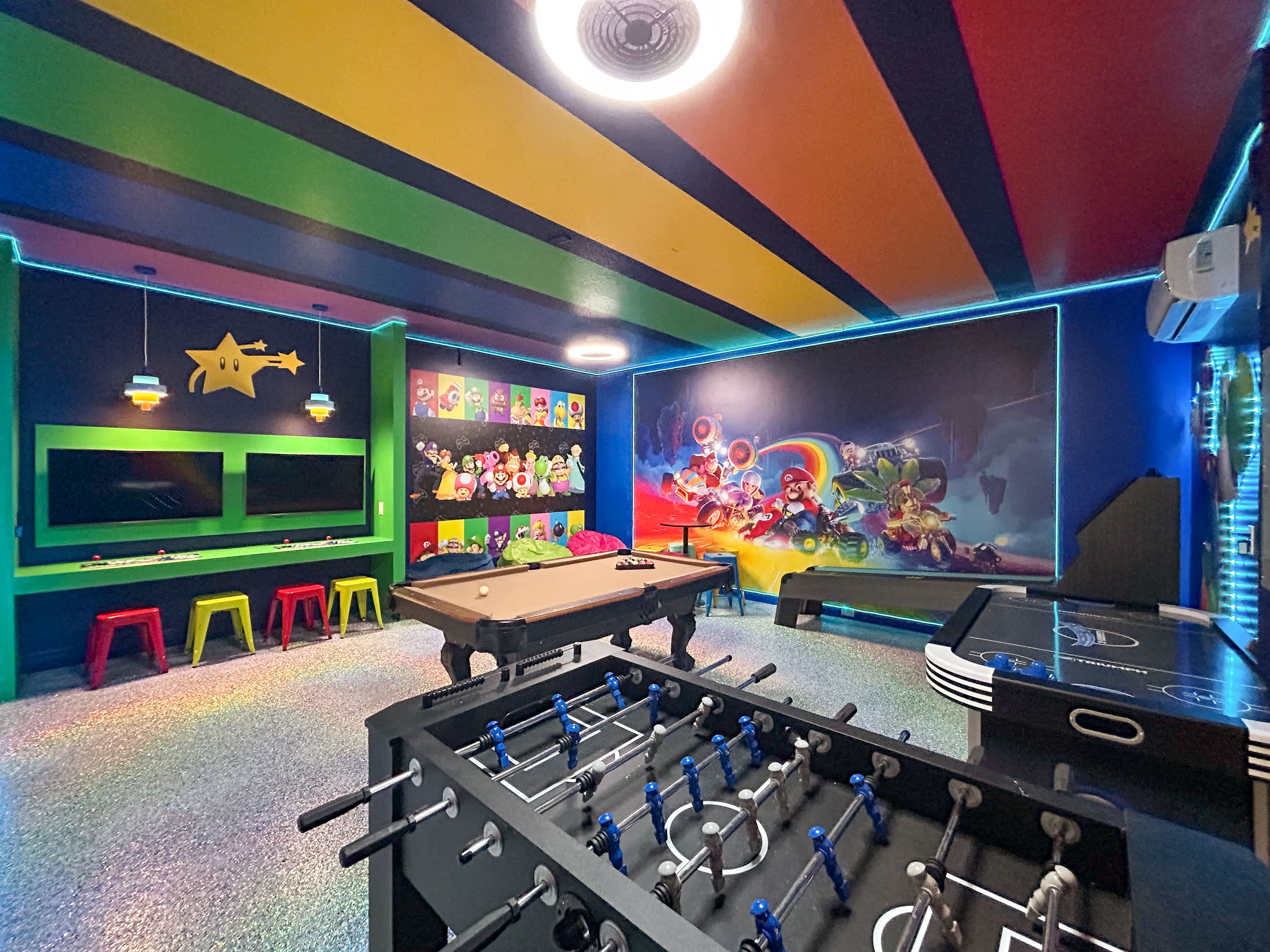 A vibrant game room featuring a pool table, foosball table, and air hockey, set against walls adorned with Mario-themed murals and bright neon lighting. The ceiling is painted with colorful stripes, and there are multiple screens for gaming.