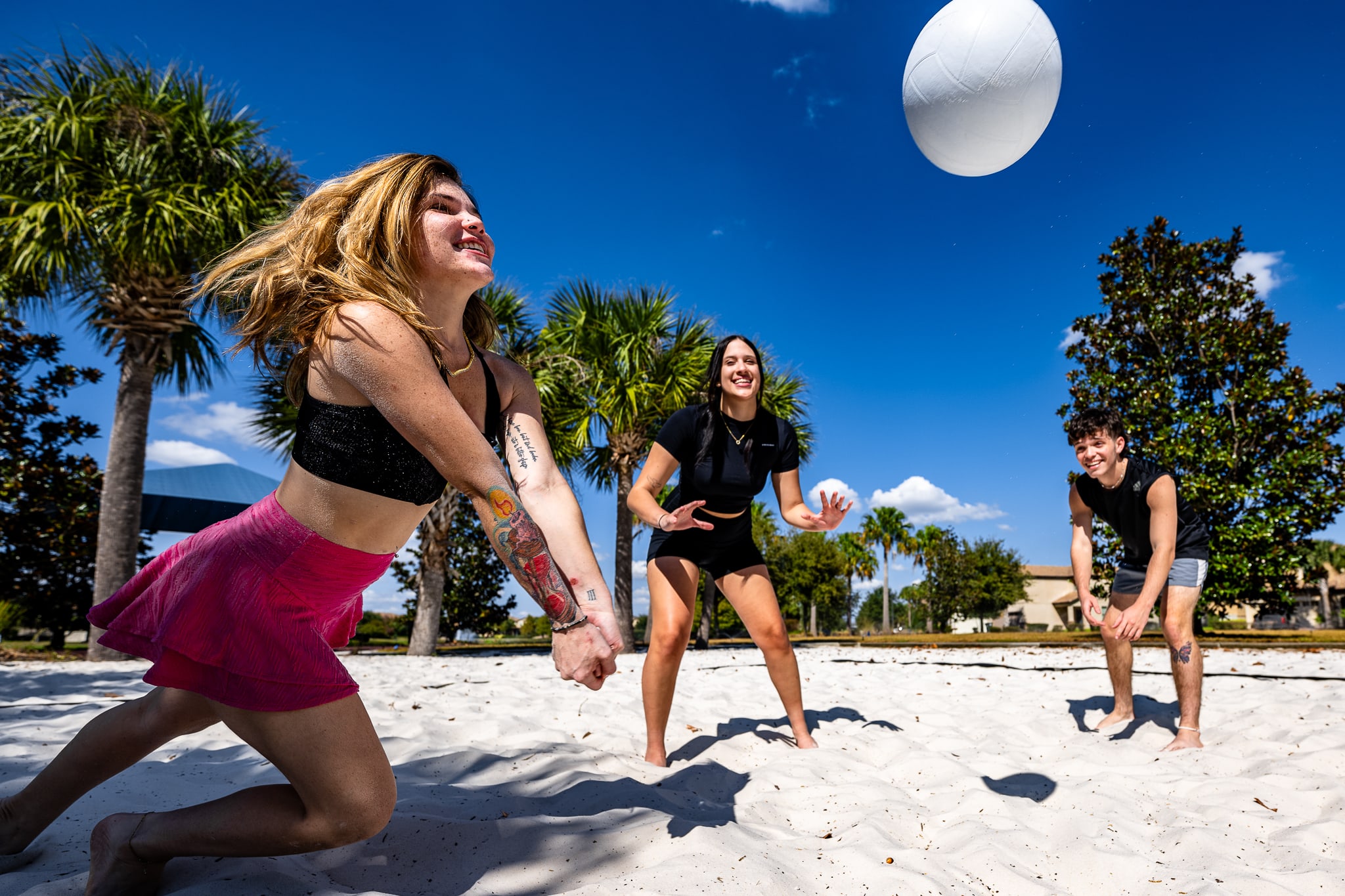 Three young adults are actively playing beach volleyball on a sandy court under a bright blue sky with palm trees in the background. One person is hitting the ball, while the others are ready to receive.