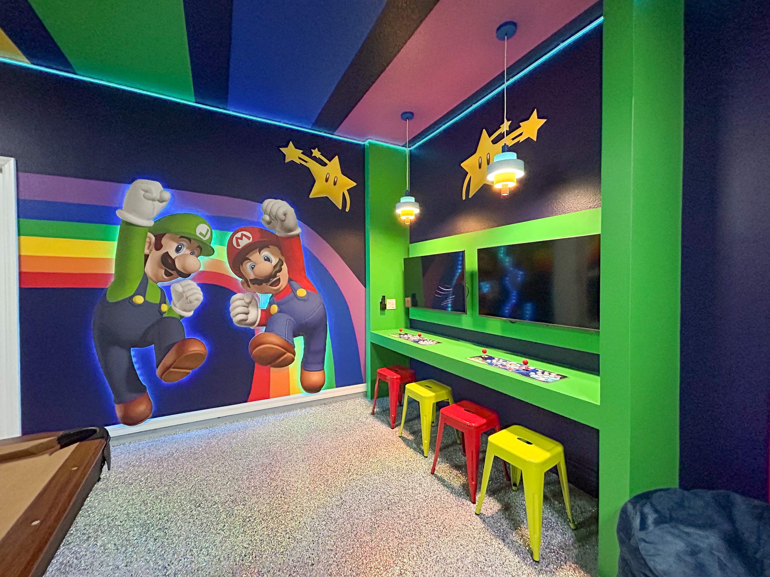 A section of the game room dedicated to video gaming, featuring two wall-mounted screens above a green console table with colorful stools. Large Mario and Luigi murals adorn the dark blue walls, complemented by neon lighting and a rainbow-striped ceiling.