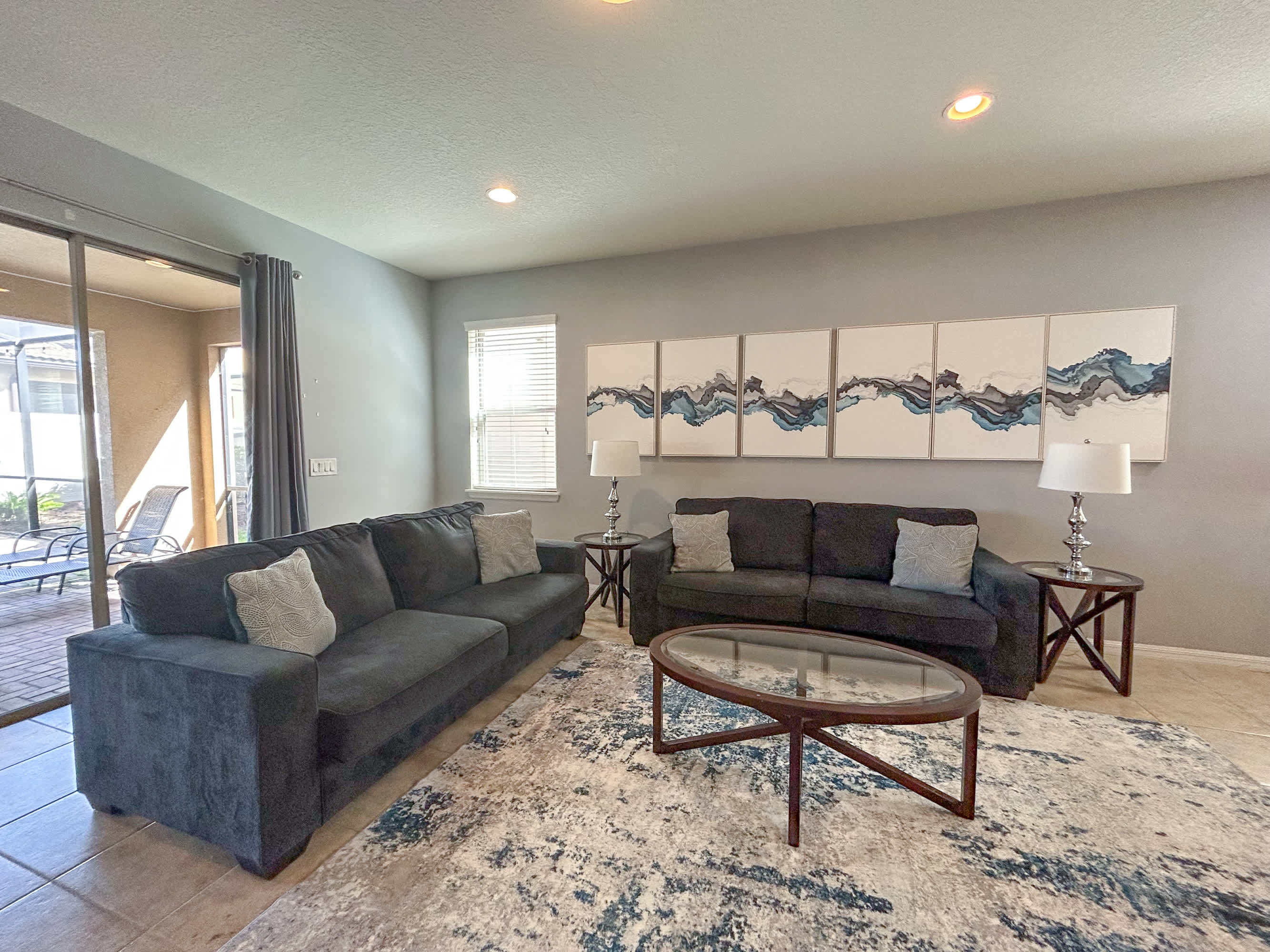 A modern living room featuring two dark gray sofas with throw pillows, a glass-top coffee table, and two end tables with lamps. The room has light gray walls, tiled floors, and a large abstract artwork above one sofa.