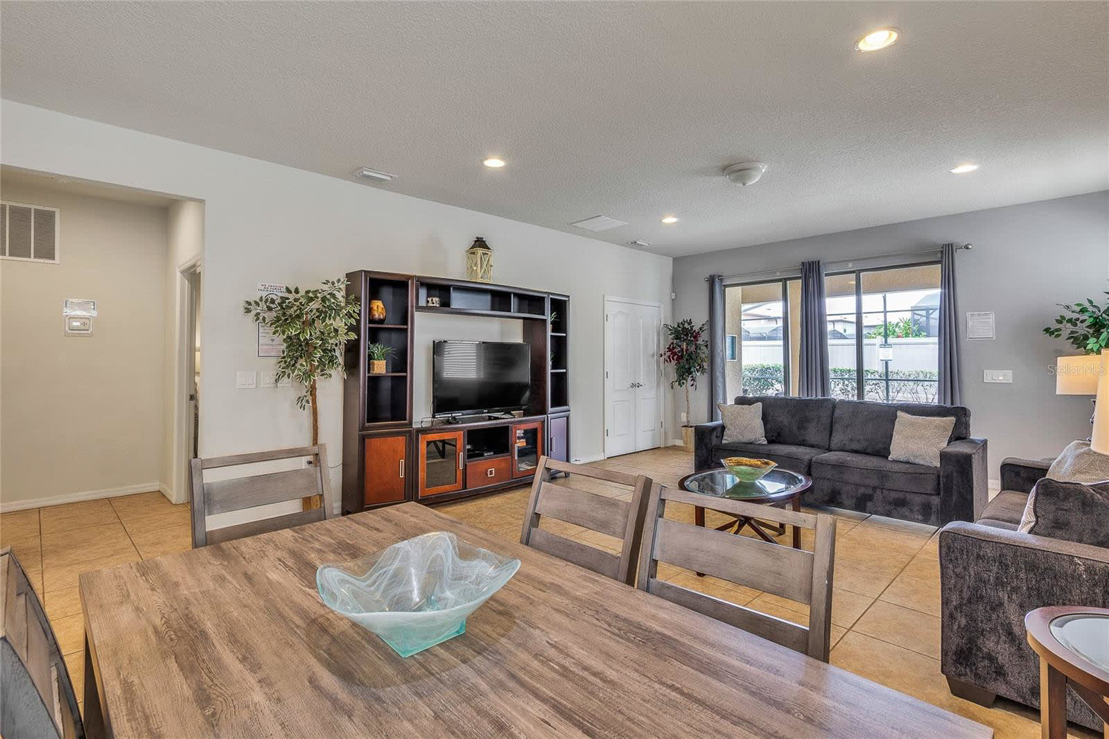 An open-concept living and dining space featuring a dark entertainment center with a TV, a dark sofa, and a rustic wooden dining table with matching chairs in the foreground.