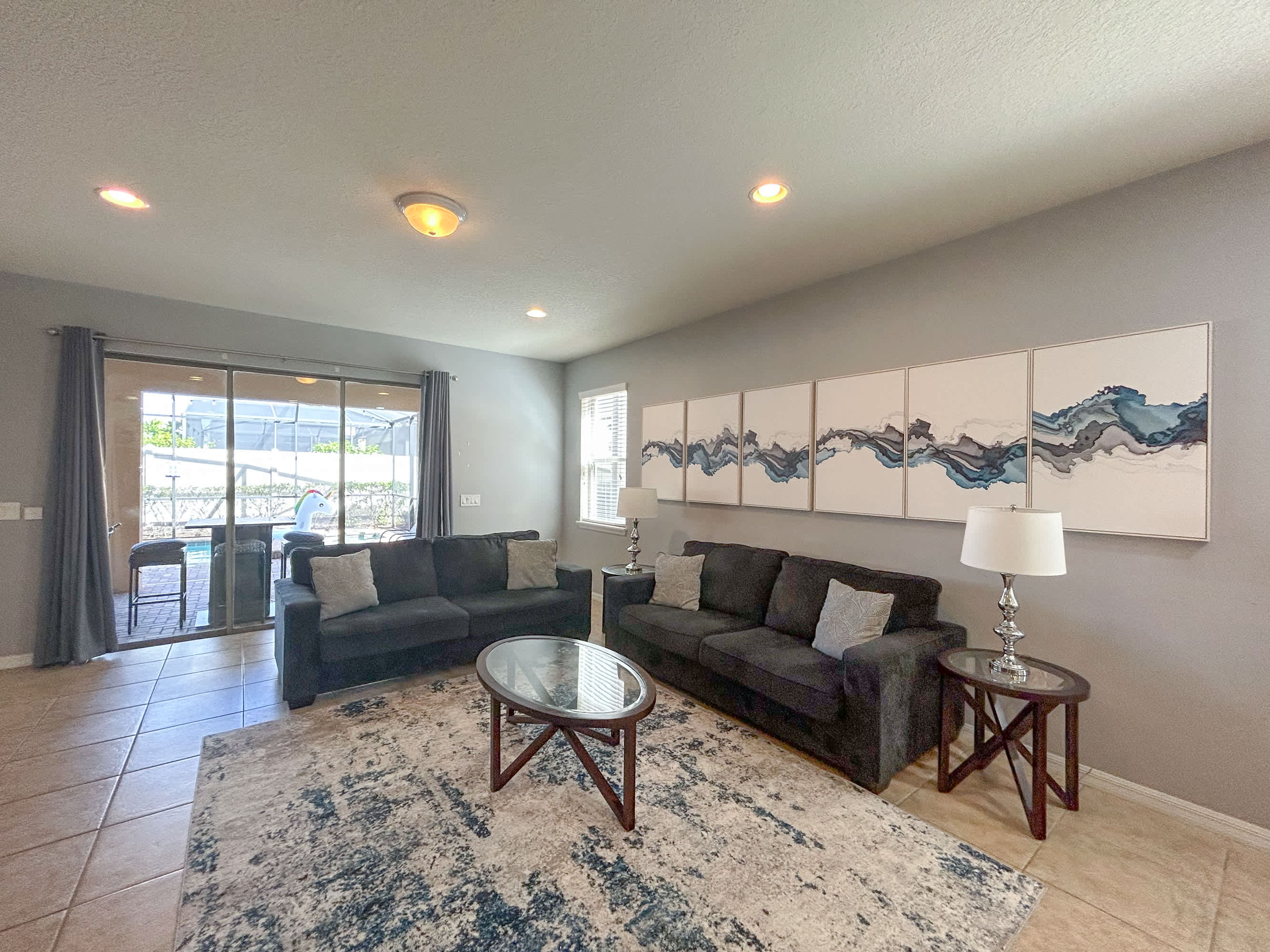 A modern living room with two dark gray sofas, a glass-top coffee table, and two end tables with lamps. The room features light gray walls, tiled floors, and a large abstract artwork above one sofa, with a view of the pool area through sliding glass doors.