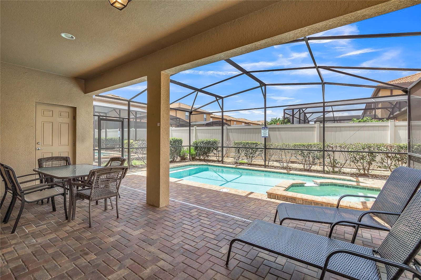A screened-in outdoor pool area with a small rectangular pool and an adjacent hot tub, surrounded by brick paving. The covered patio features an outdoor dining set and two lounge chairs, with a privacy fence in the background.