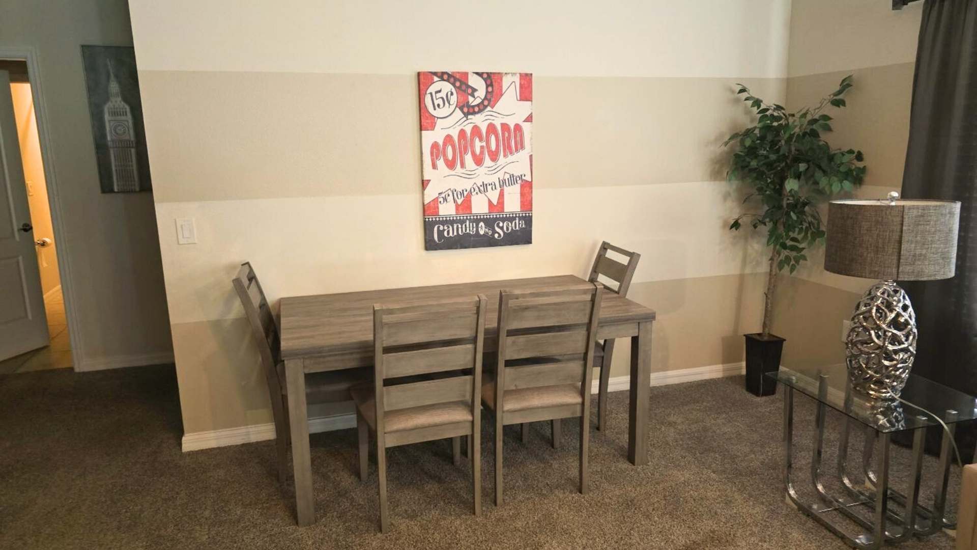 A casual dining area with a rustic wooden table and four matching chairs with upholstered seats, set on carpeted flooring, with a 'Popcorn' themed wall art.