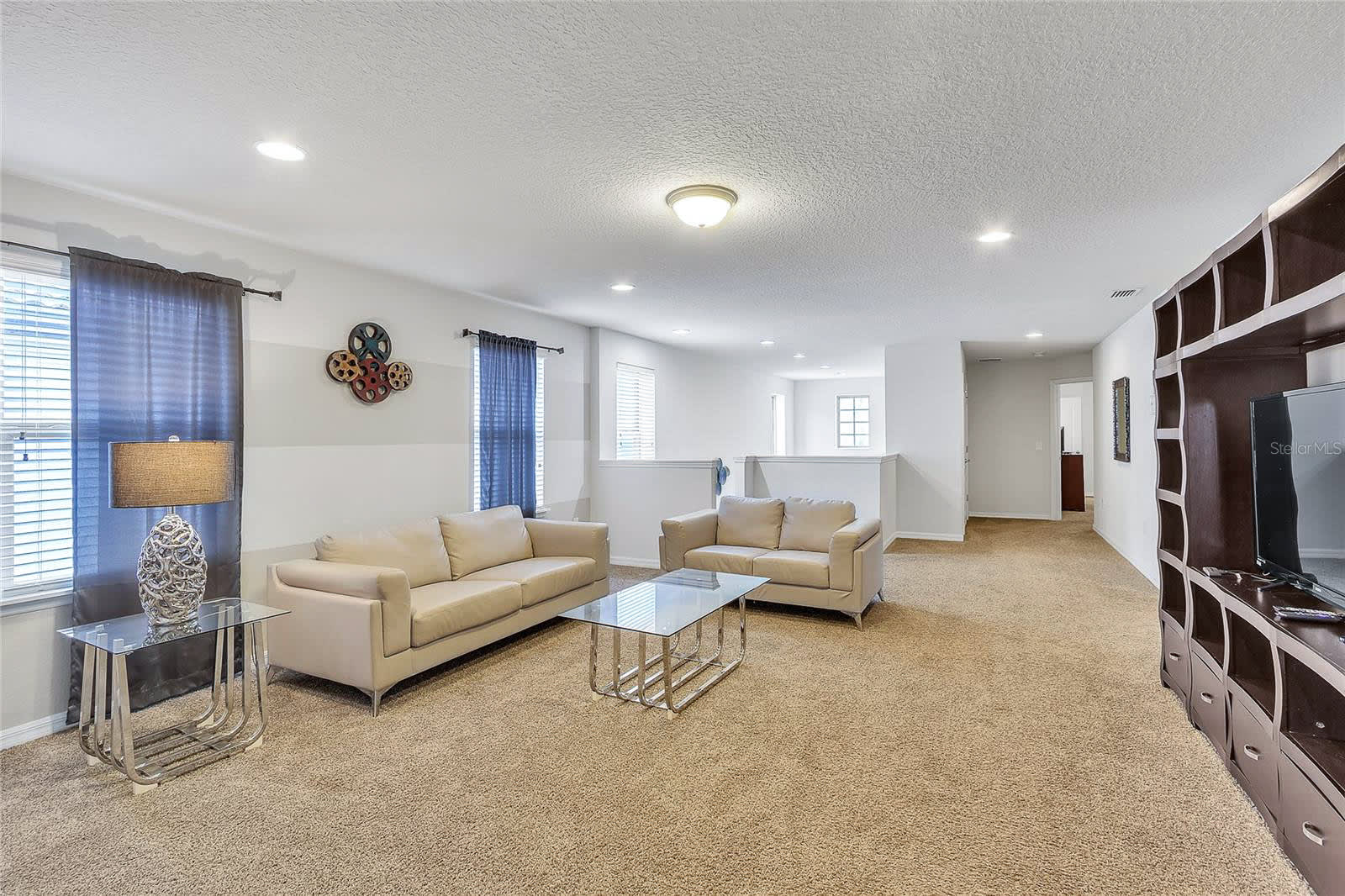 A spacious upstairs game room or loft with light beige carpet, two cream-colored sofas, a glass coffee table, and a dark wood entertainment unit with a TV. The walls are light-colored with a textured ceiling, and there's a decorative wall hanging with film reels.