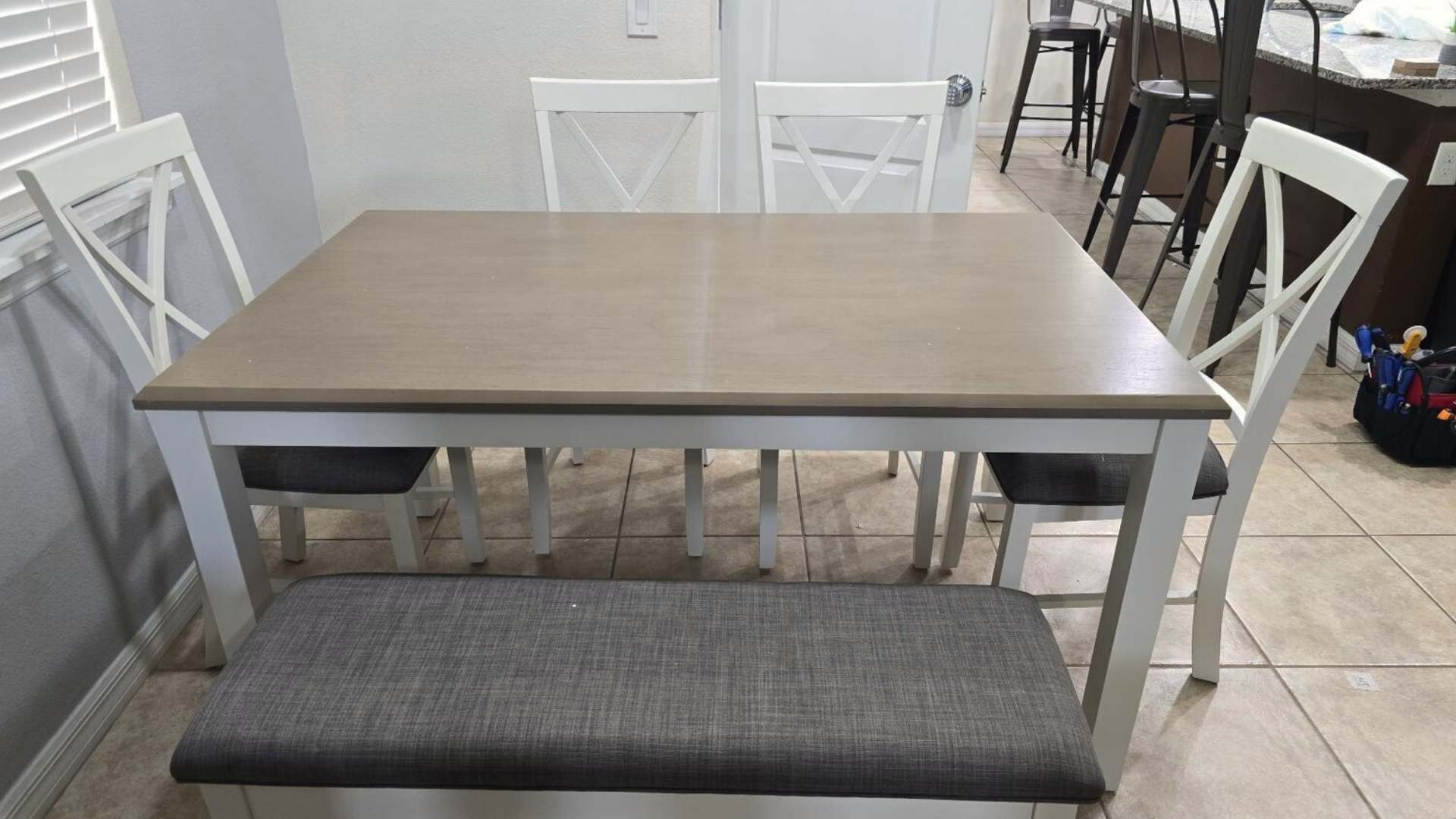 A modern dining area featuring a light wood-top table with white legs, accompanied by white cross-back chairs and a grey upholstered bench, set against a light-colored wall.