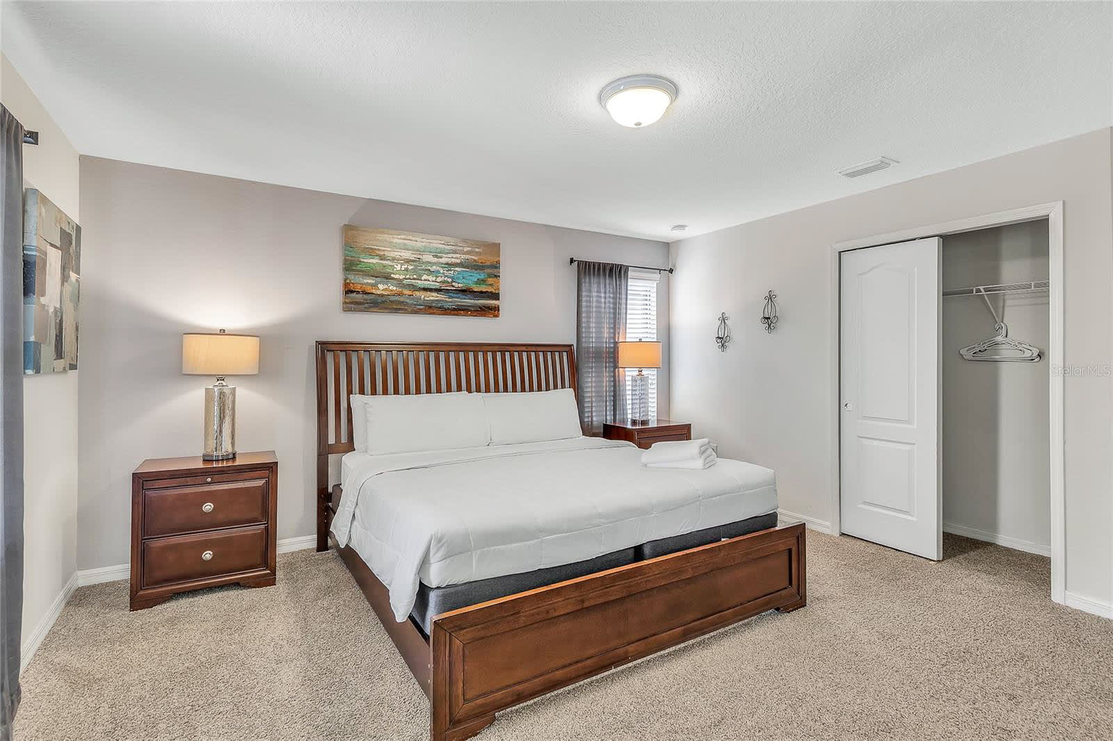 A cozy bedroom featuring a dark wood king-sized bed with white bedding, two matching nightstands with lamps, and a large abstract painting above the headboard. A sliding closet door is visible on the right, and the room has light gray walls and carpeted flooring.