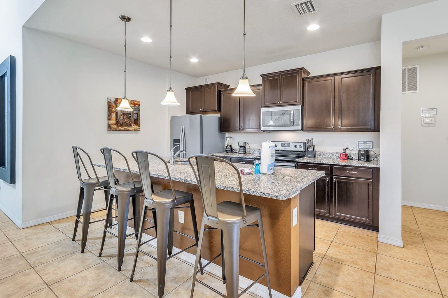 A well-lit kitchen featuring dark wood cabinetry, granite countertops, a stainless steel refrigerator, and a large island with four metal bar stools, illuminated by pendant lights.
