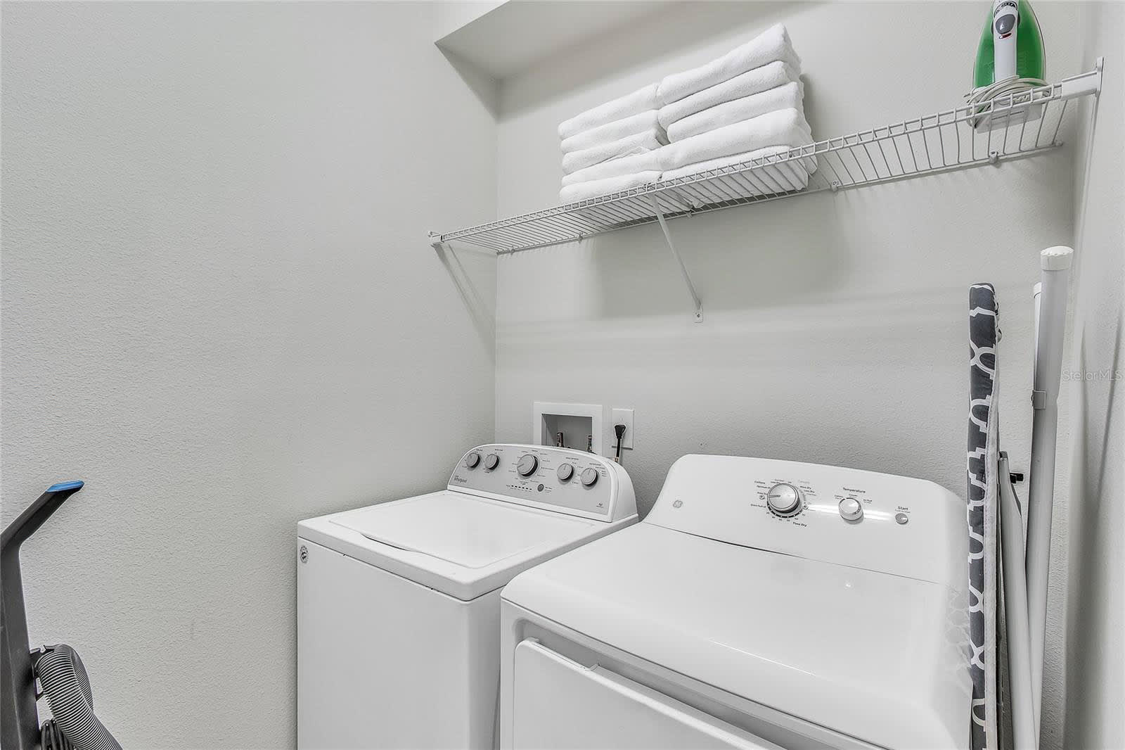 A functional laundry room equipped with a white top-loading washing machine and a front-loading dryer, positioned beneath a wire shelf stocked with neatly folded white towels. An ironing board and iron are also visible, indicating full laundry facilities.