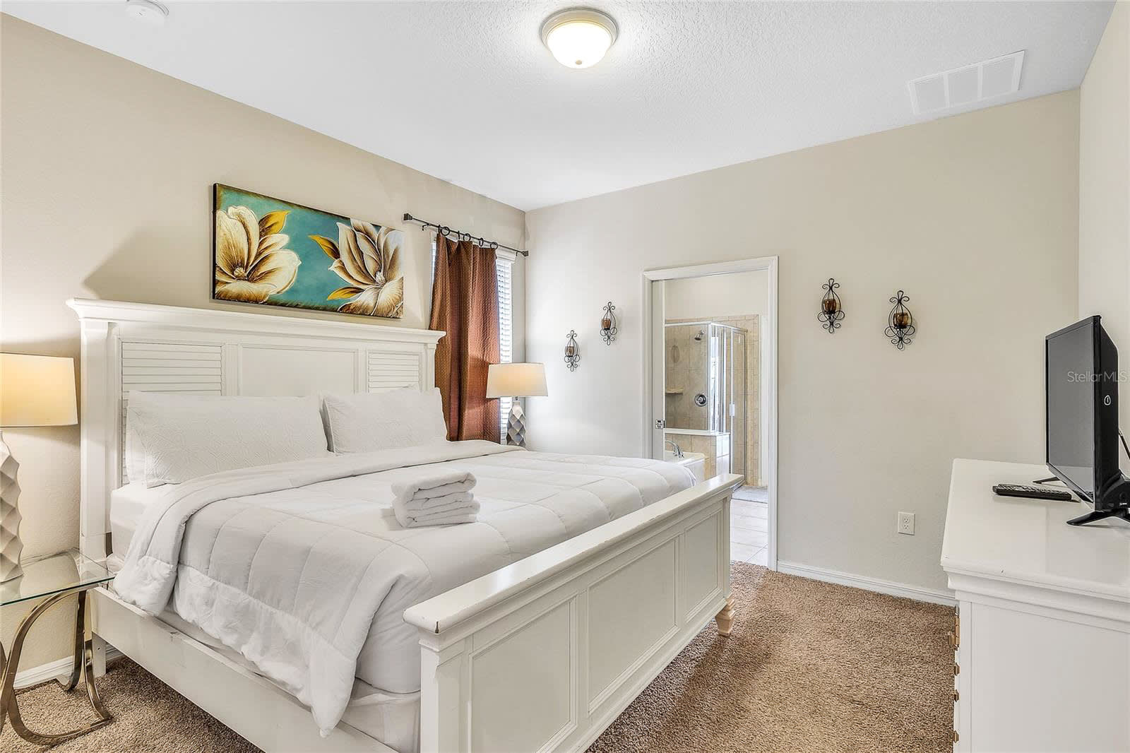 A bright bedroom with a white slatted headboard bed, white bedding, a white dresser with a TV, and a glass-top nightstand, featuring light brown carpet and a large floral artwork.