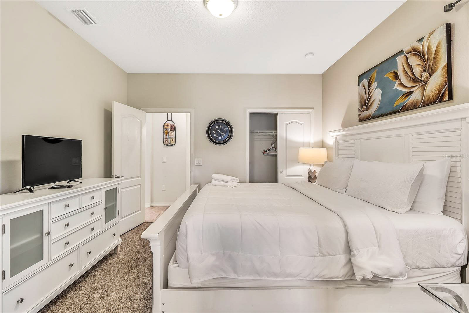 A cozy bedroom featuring a white slatted bed with white linens, a matching white dresser with a TV, and a closet, all set on light brown carpet with a large floral painting above the bed.