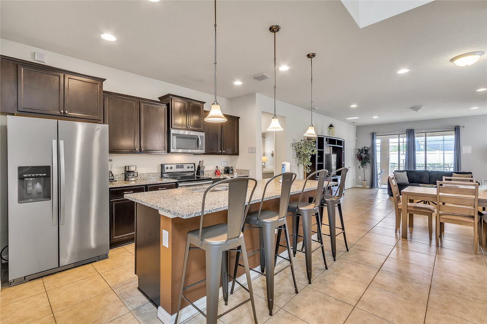 A spacious open-concept kitchen with dark wood cabinets, granite countertops, stainless steel appliances, and a large island with four industrial-style bar stools, leading to a dining area.