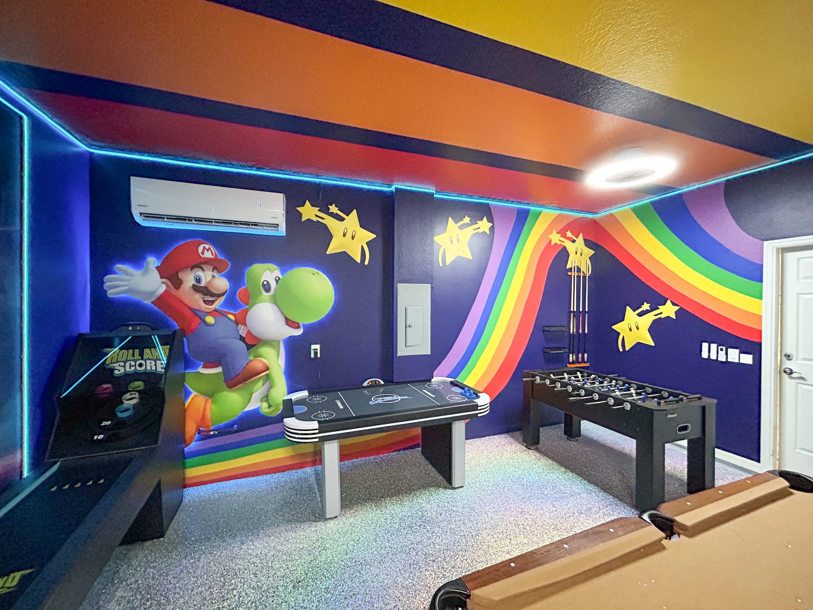 A brightly lit game room featuring a foosball table, an air hockey table, and a pool table, with Mario and Yoshi murals on the dark blue walls. The ceiling is painted with colorful stripes, and neon lights illuminate the space.