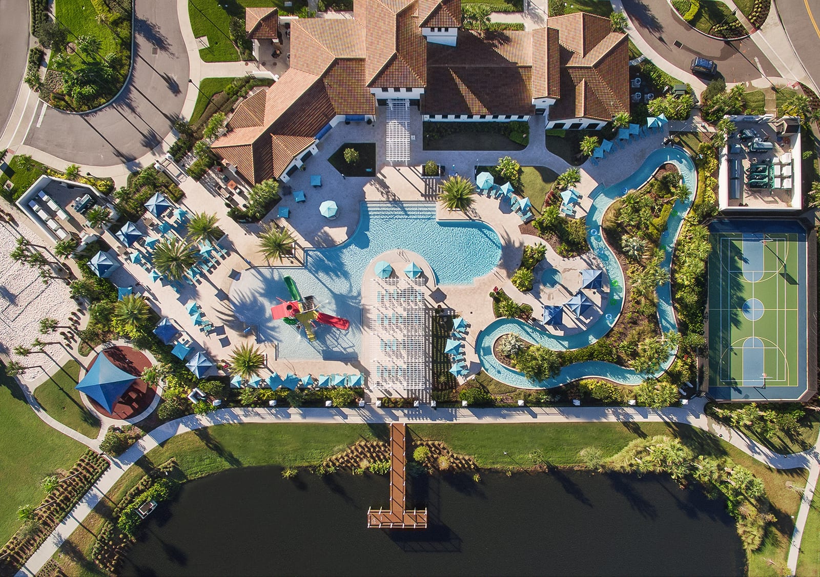 A detailed aerial view showcasing the resort's main pool area with multiple swimming pools, a vibrant blue lazy river, and a nearby outdoor basketball court. Lush green landscaping and palm trees surround the amenities.