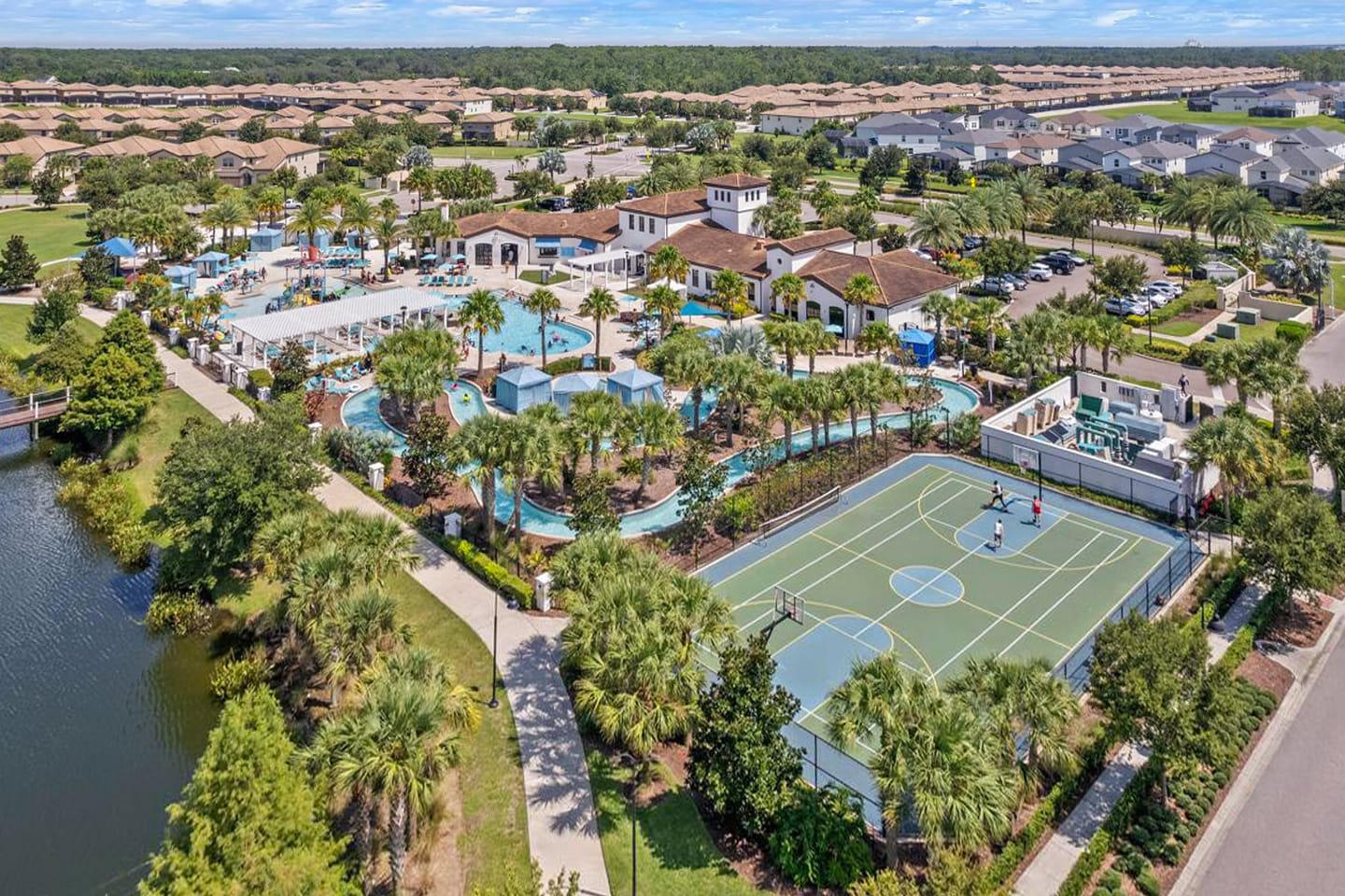 An expansive aerial view of a resort community featuring a large central clubhouse, multiple swimming pools, a winding lazy river, and a full-size outdoor basketball court. Numerous residential homes are visible in the surrounding landscape.