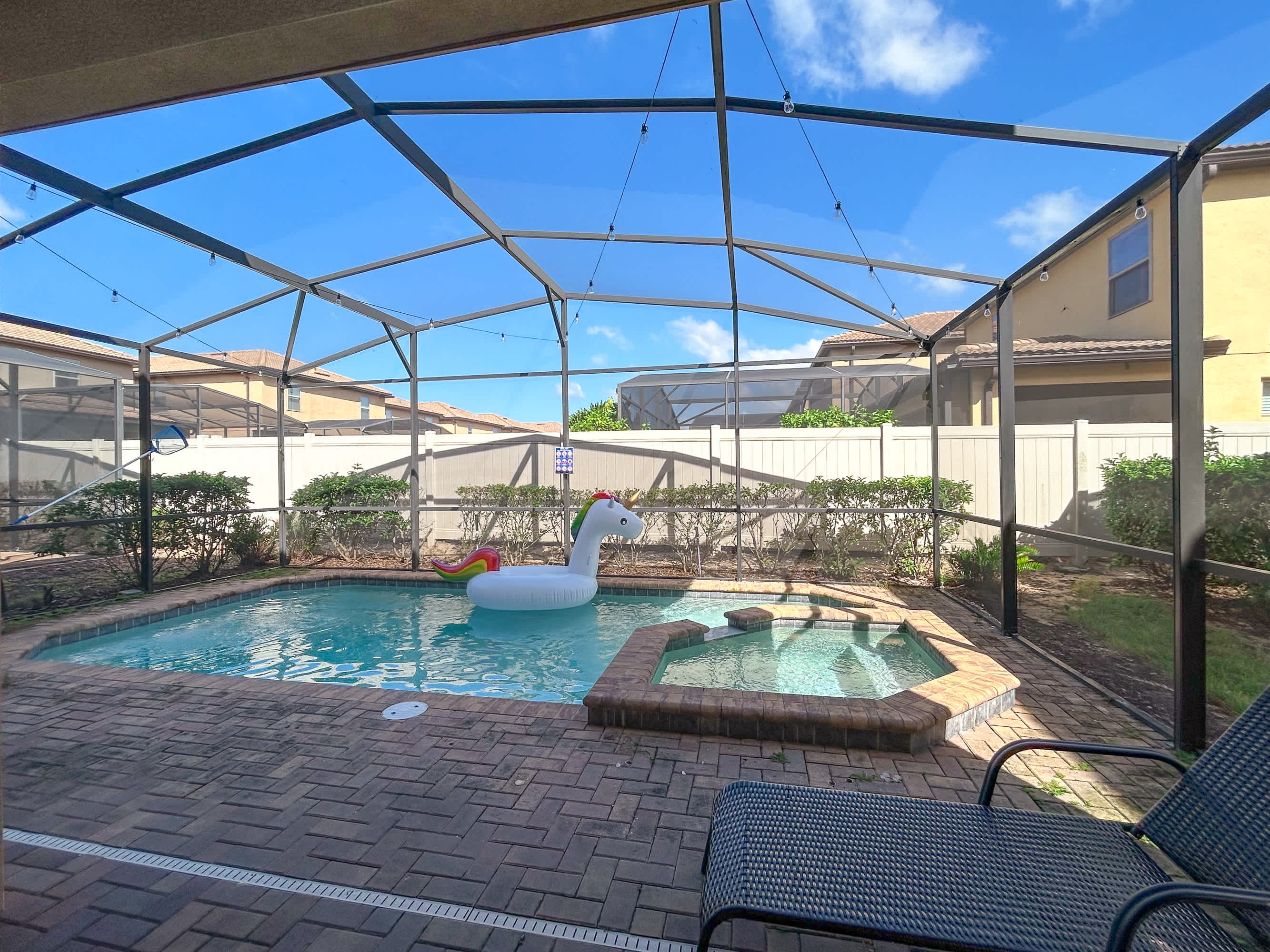 A private screened-in swimming pool with a built-in hot tub, featuring a unicorn float and brick paver decking. Two lounge chairs are visible, offering a relaxing outdoor space.