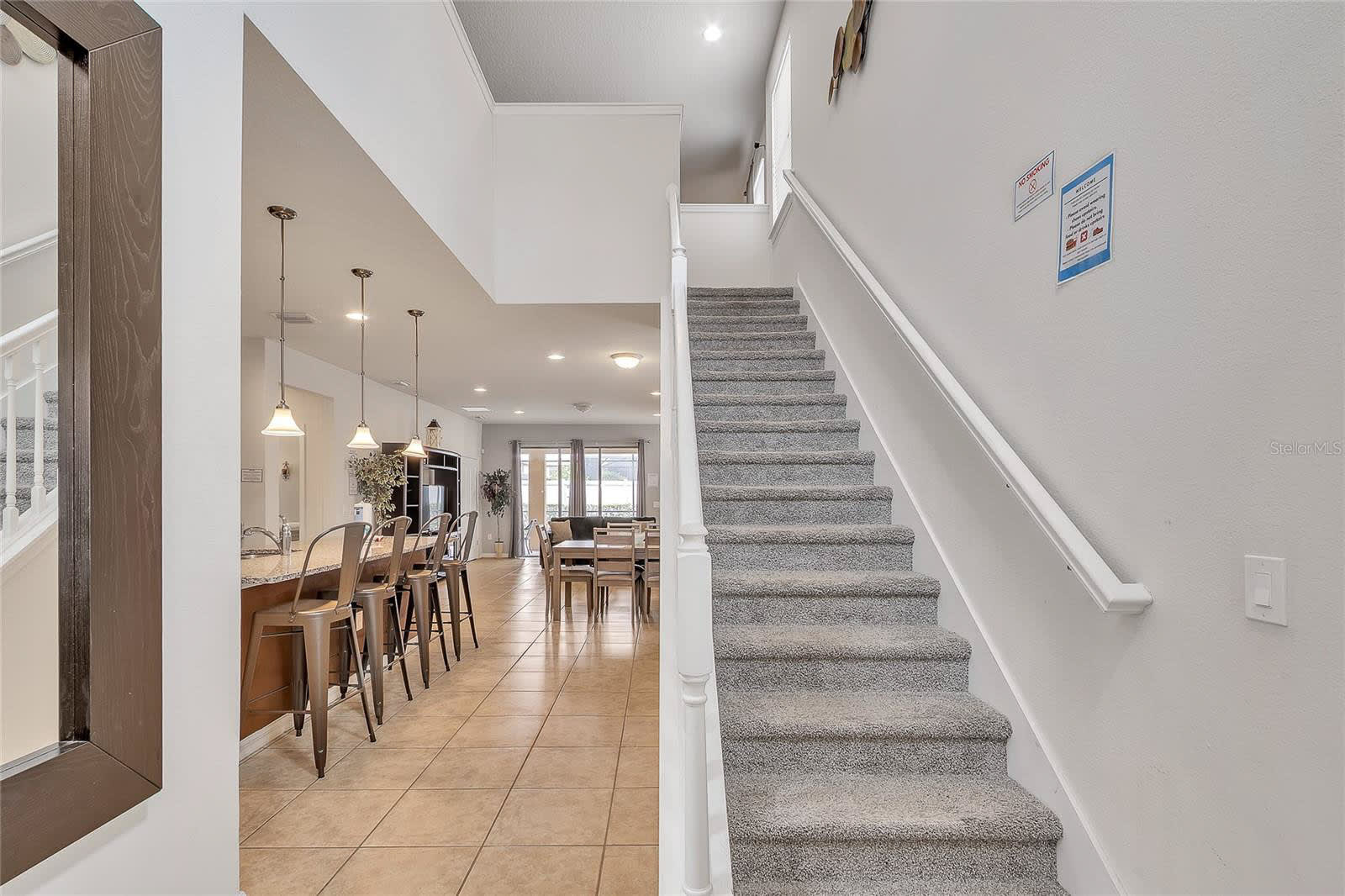 A bright entryway featuring a carpeted staircase with white railings leading upstairs, offering an open view into the kitchen and living areas with tile flooring.