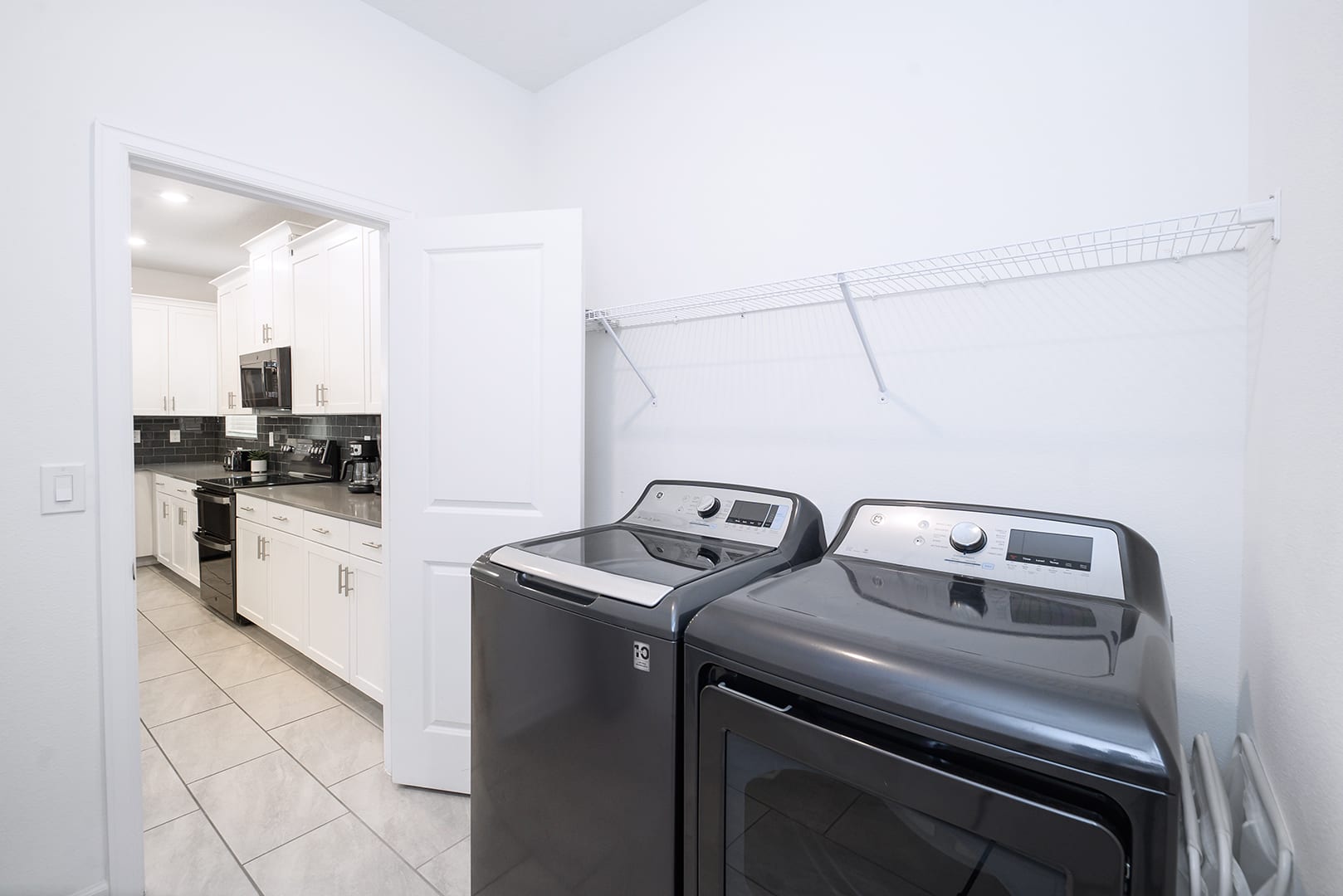 Functional laundry room equipped with a dark gray top-load washing machine and a matching front-load dryer. A wire shelf is mounted above, and a doorway leads into a kitchen area.