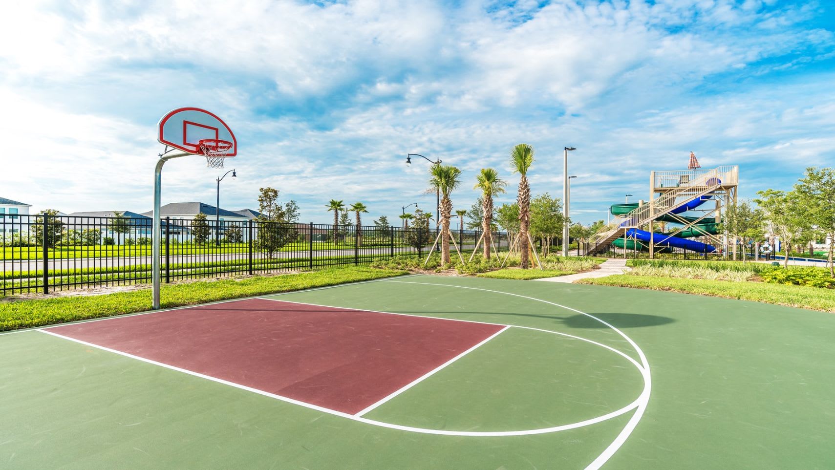 An outdoor basketball court with a green and maroon playing surface and a red hoop stands under a bright blue sky. In the background, palm trees and the communal water slide structure are visible, suggesting a resort-style community.