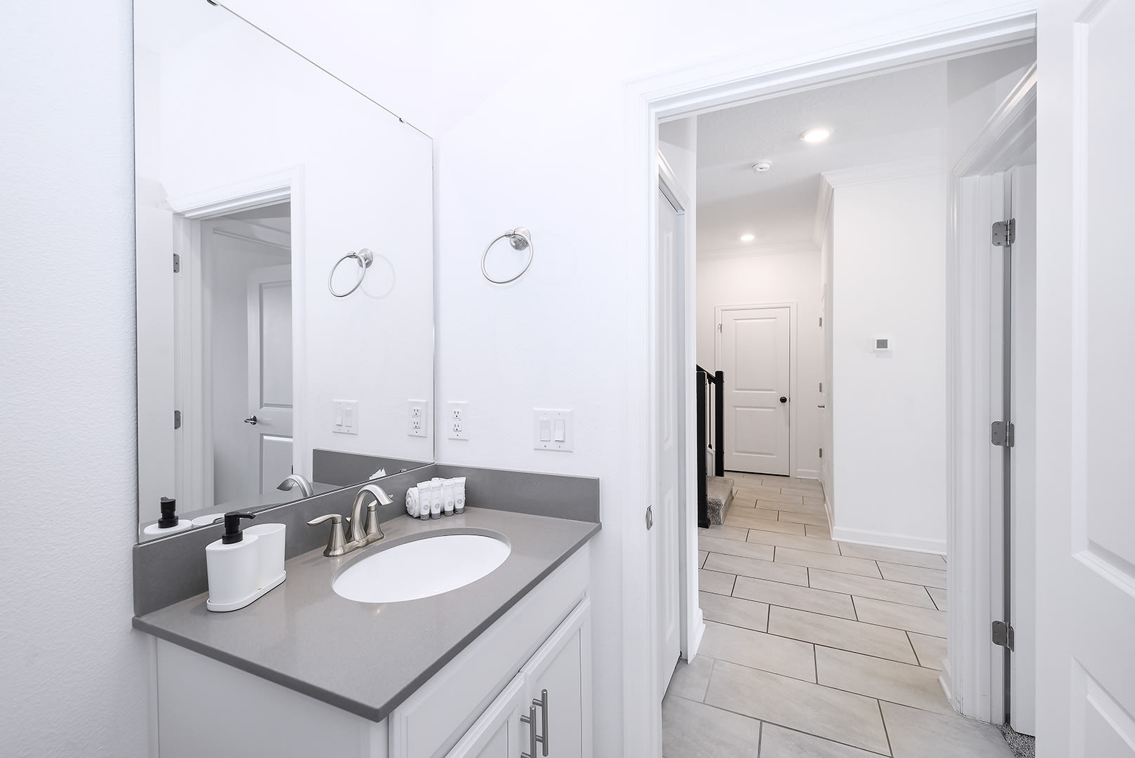 Modern bathroom vanity area featuring a single sink set into a gray countertop, a large frameless mirror, and white cabinetry. Toiletries are neatly arranged on the counter, with a hallway visible through the doorway.
