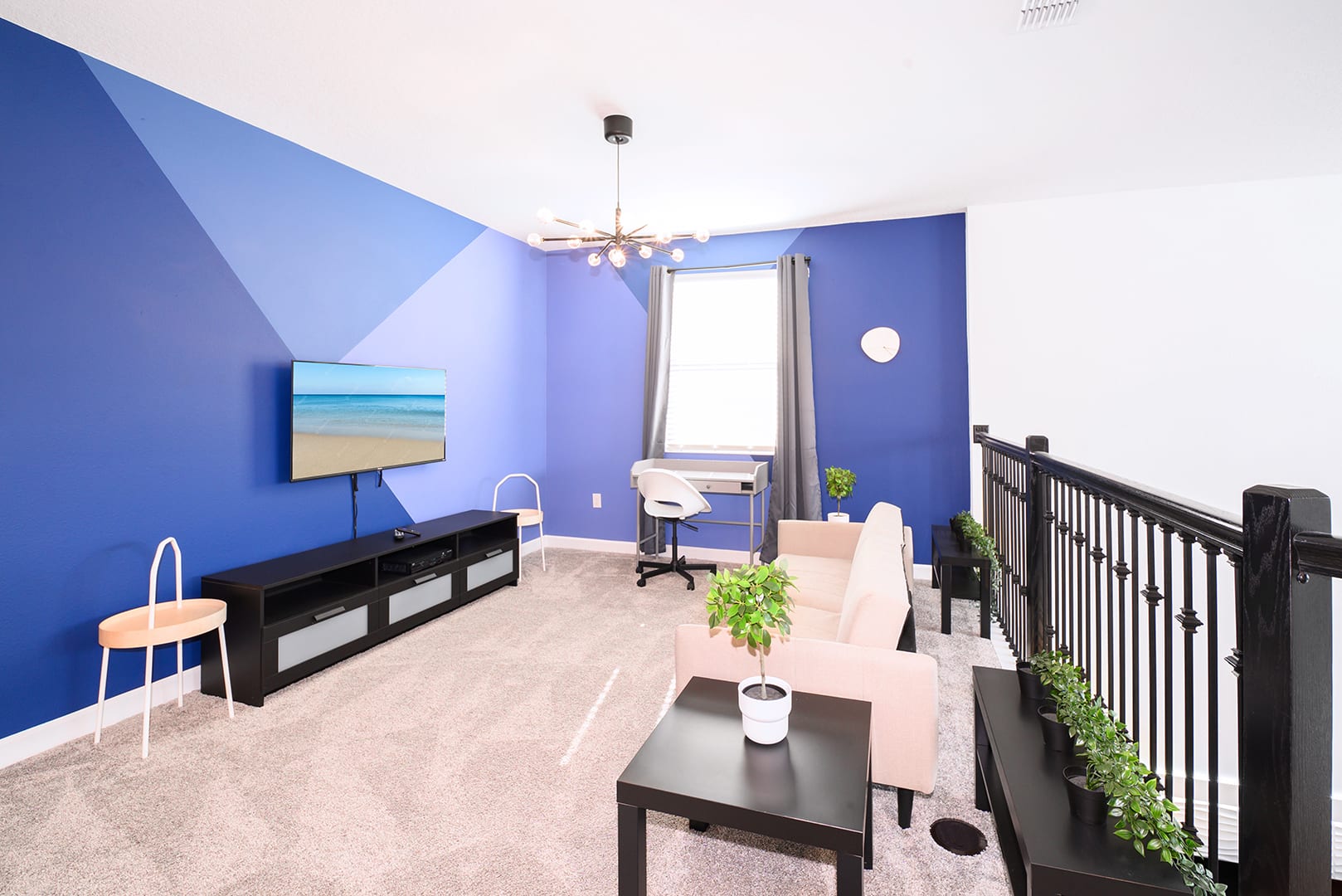An upstairs loft area features a large flat-screen TV on a black console against a blue geometric accent wall, a light pink sofa, a desk with a chair, and carpeted floors.