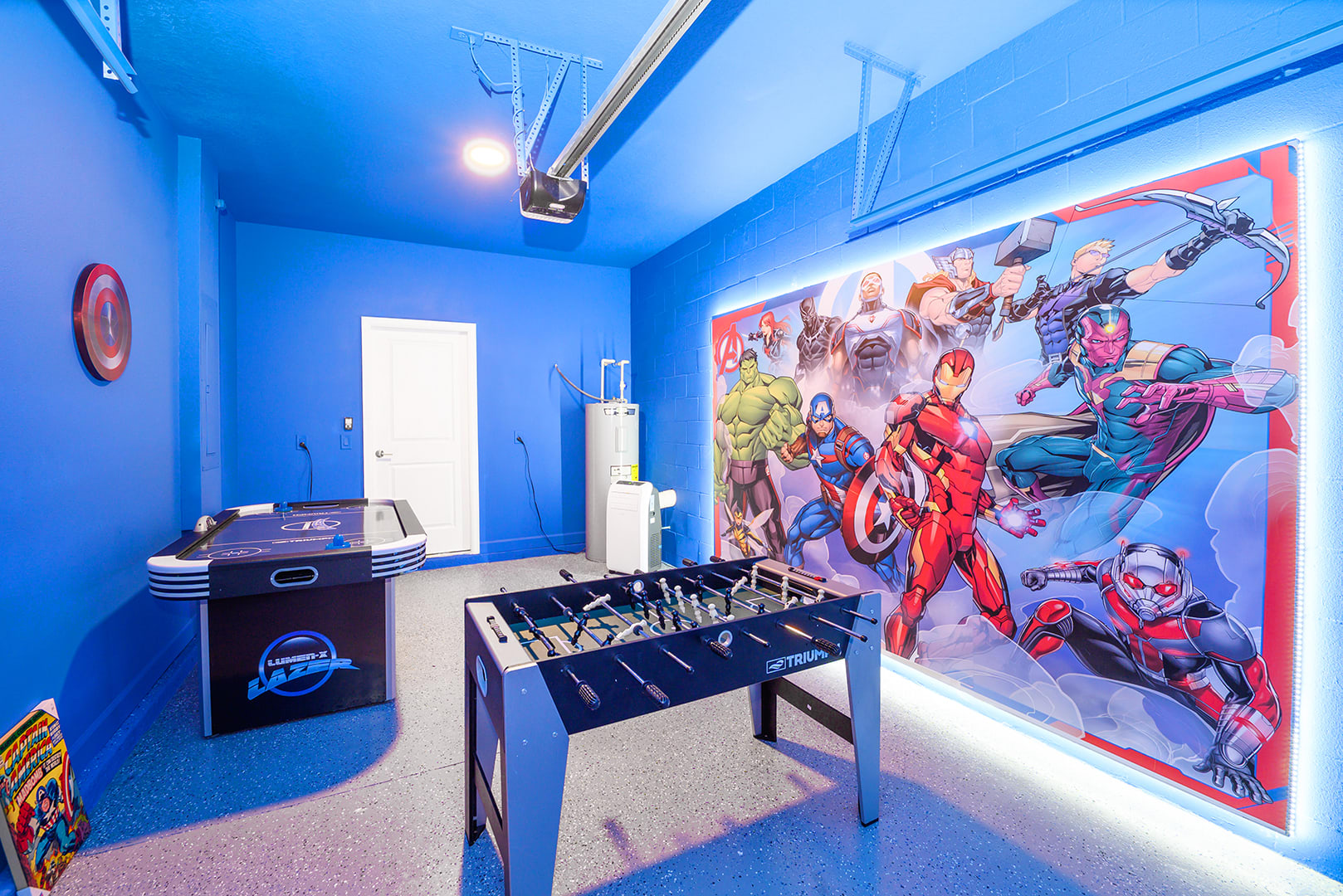A vibrant blue-painted garage converted into a game room features a foosball table, an air hockey table, and a large backlit Marvel superhero mural on one wall, with a speckled epoxy floor.
